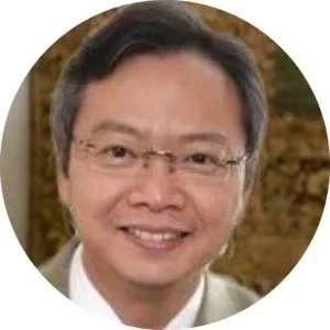 A close-up portrait of an Asian man with glasses, smiling, with a blurred outdoor background.