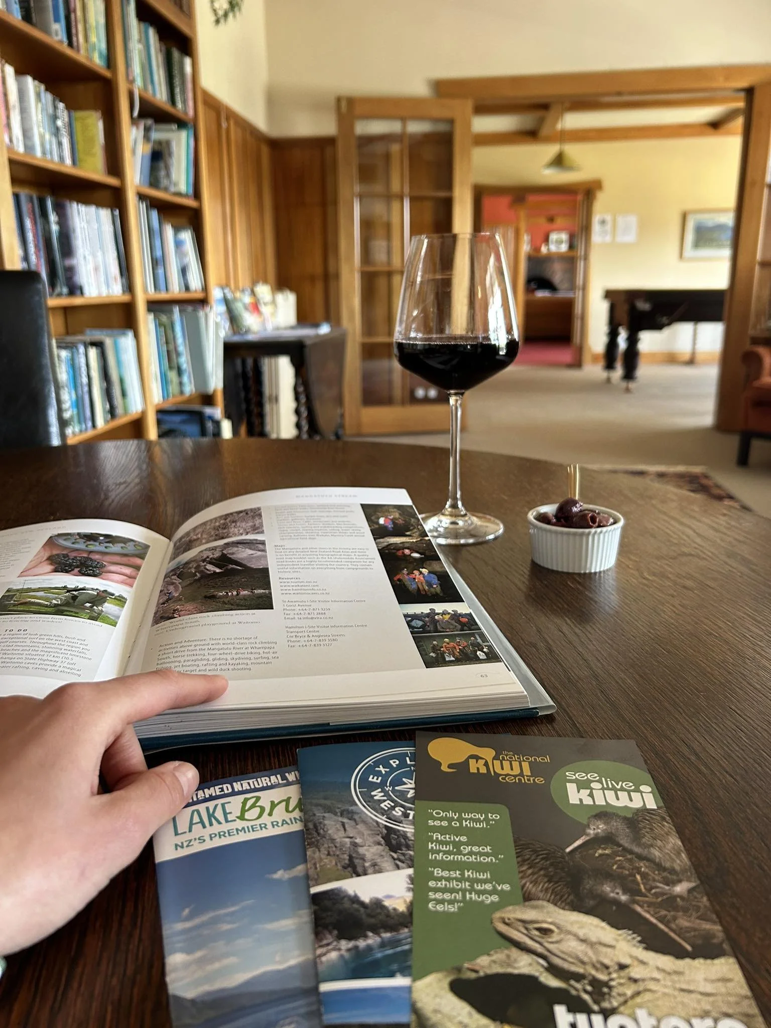 A relaxing moment in the library at Lake Brunner Eco Lodge, with a glass of red wine, an open book and local West Coast visitor guides on a wooden table beside bookshelves.