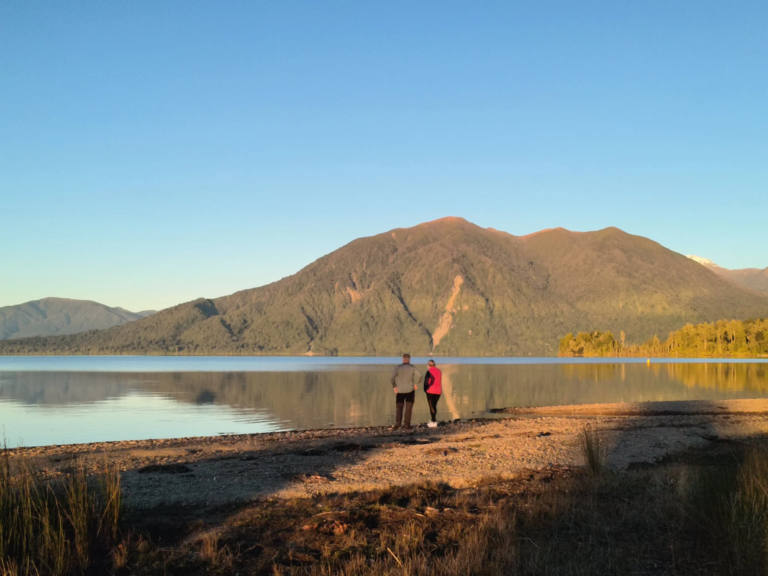 Couple standing beside Lake Brunner with  mountain reflections across the water on New Zealand's West Coast.