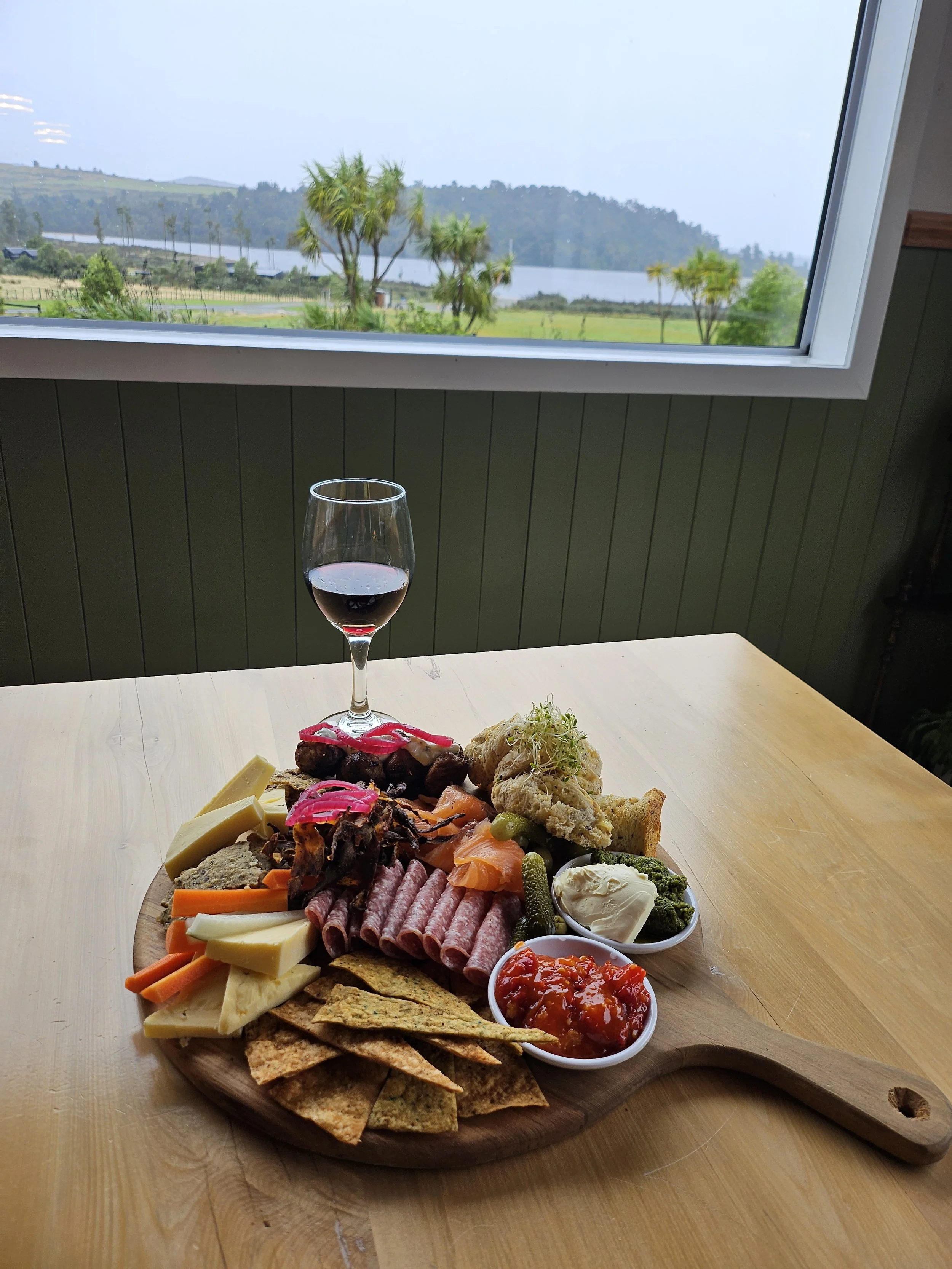 Charcuterie board with wine overlooking Lake Brunner on New Zealand’s West Coast at Lake Brunner Eco Lodge. Hosted eco-lodge dining experience featuring local food and peaceful lake views,