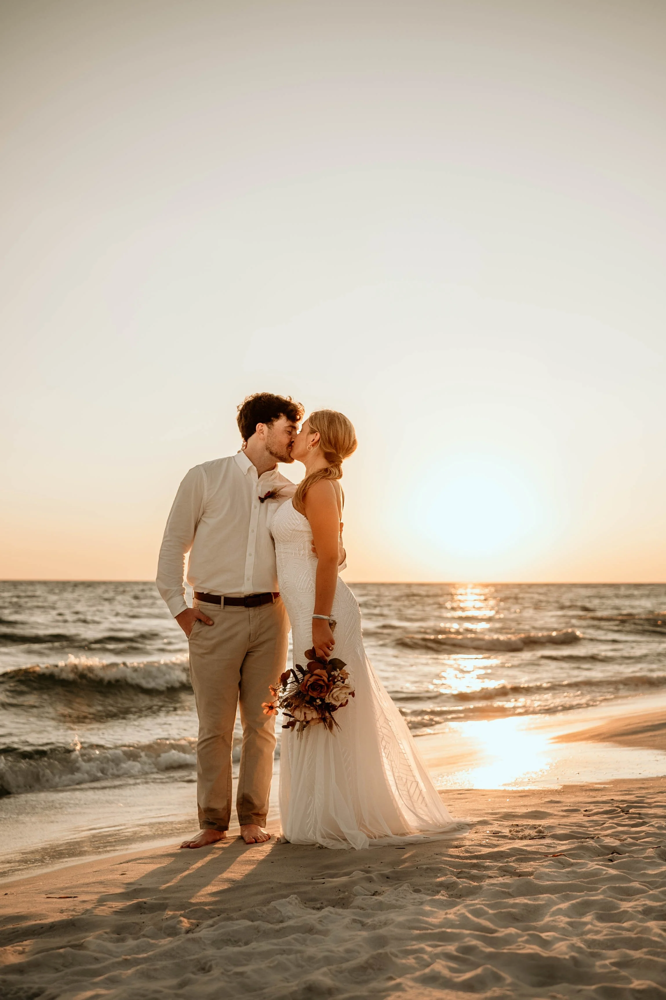A couple in wedding attire sharing a kiss on the beach during sunset, with the ocean waves in the background. Panama City beach weddings, PCB Photographers, PCB weddings