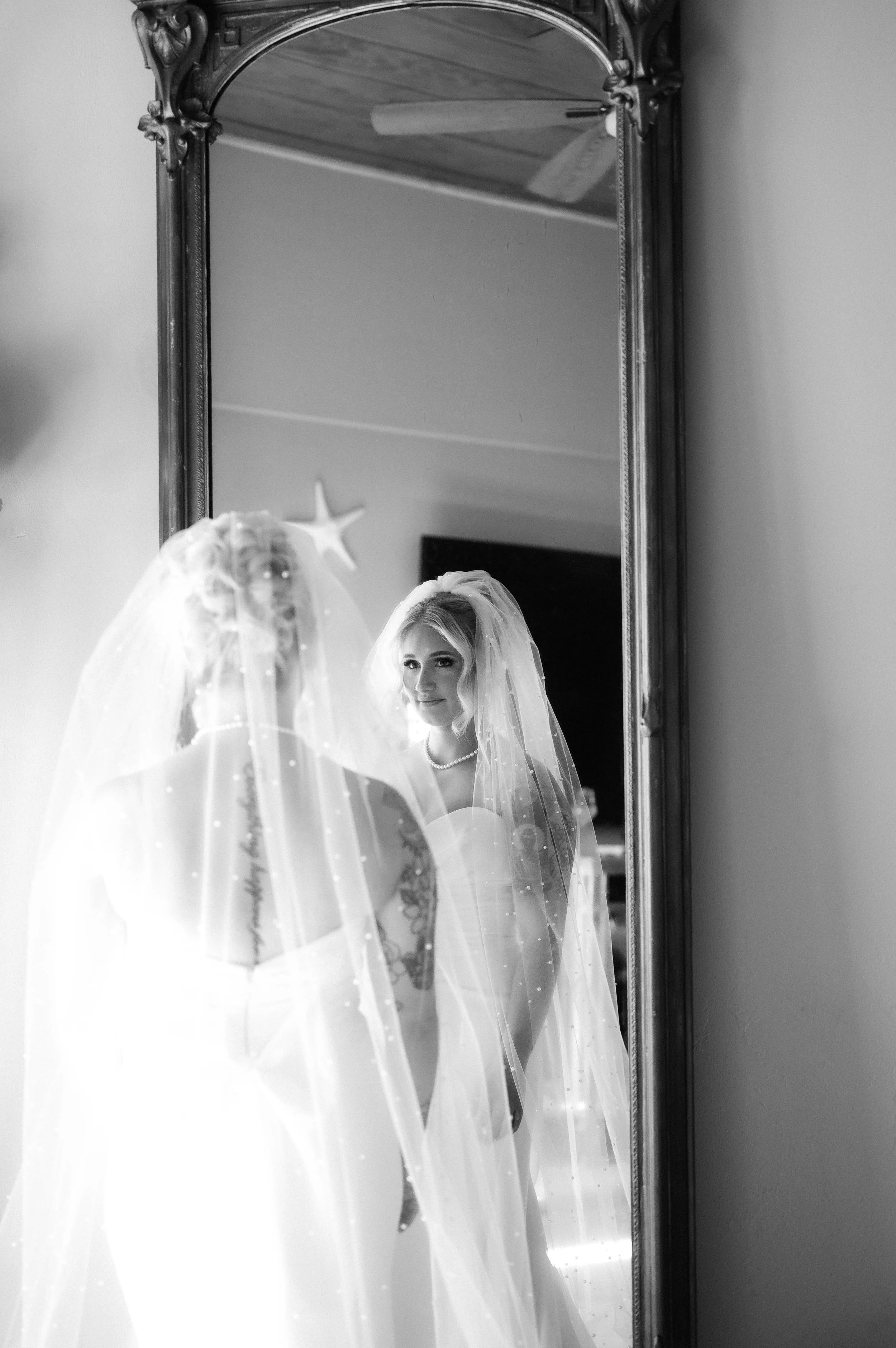 Black and white photo of a bride looking at her reflection in a tall mirror, wearing a wedding dress, veil, and pearl necklace. Panama City beach wedding photographer