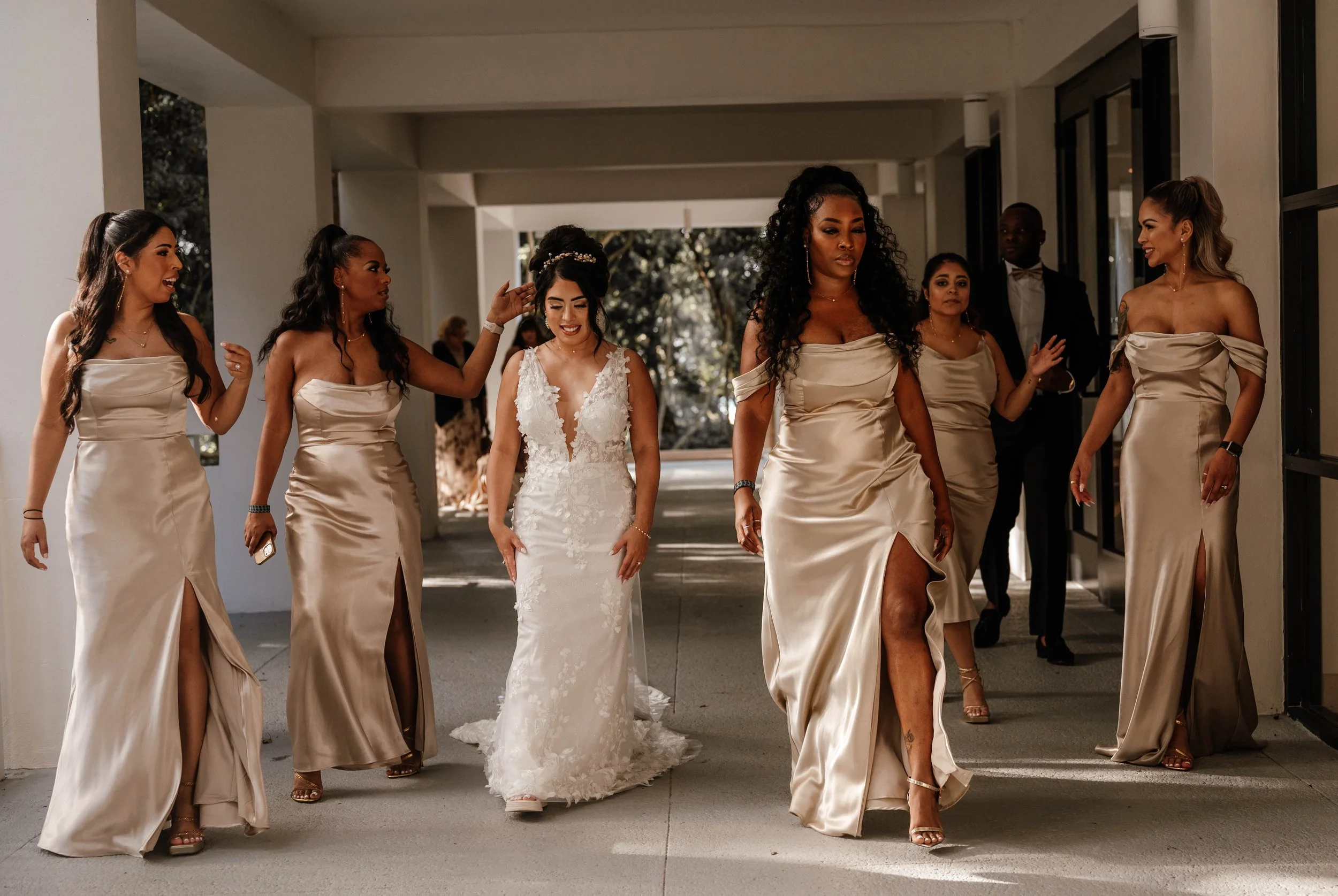 A bride and six bridesmaids walking together in a corridor, all dressed in elegant satin dresses, with the bride in a white lace gown and the bridesmaids in matching champagne-colored gowns.