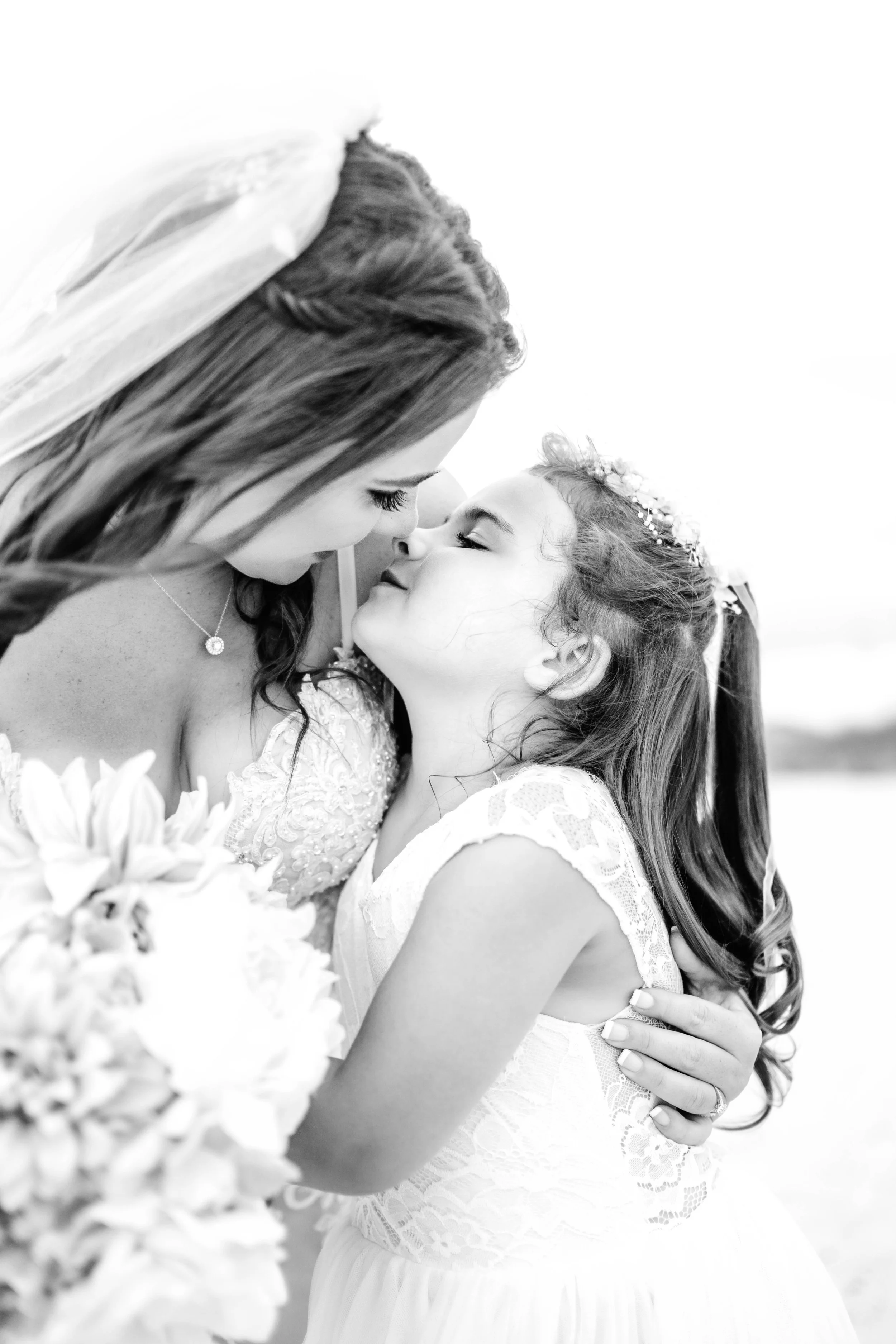 A black and white photo of a woman and a young girl, likely a bride and flower girl, sharing an intimate moment with their foreheads touching, both smiling softly. pcb wedding photography