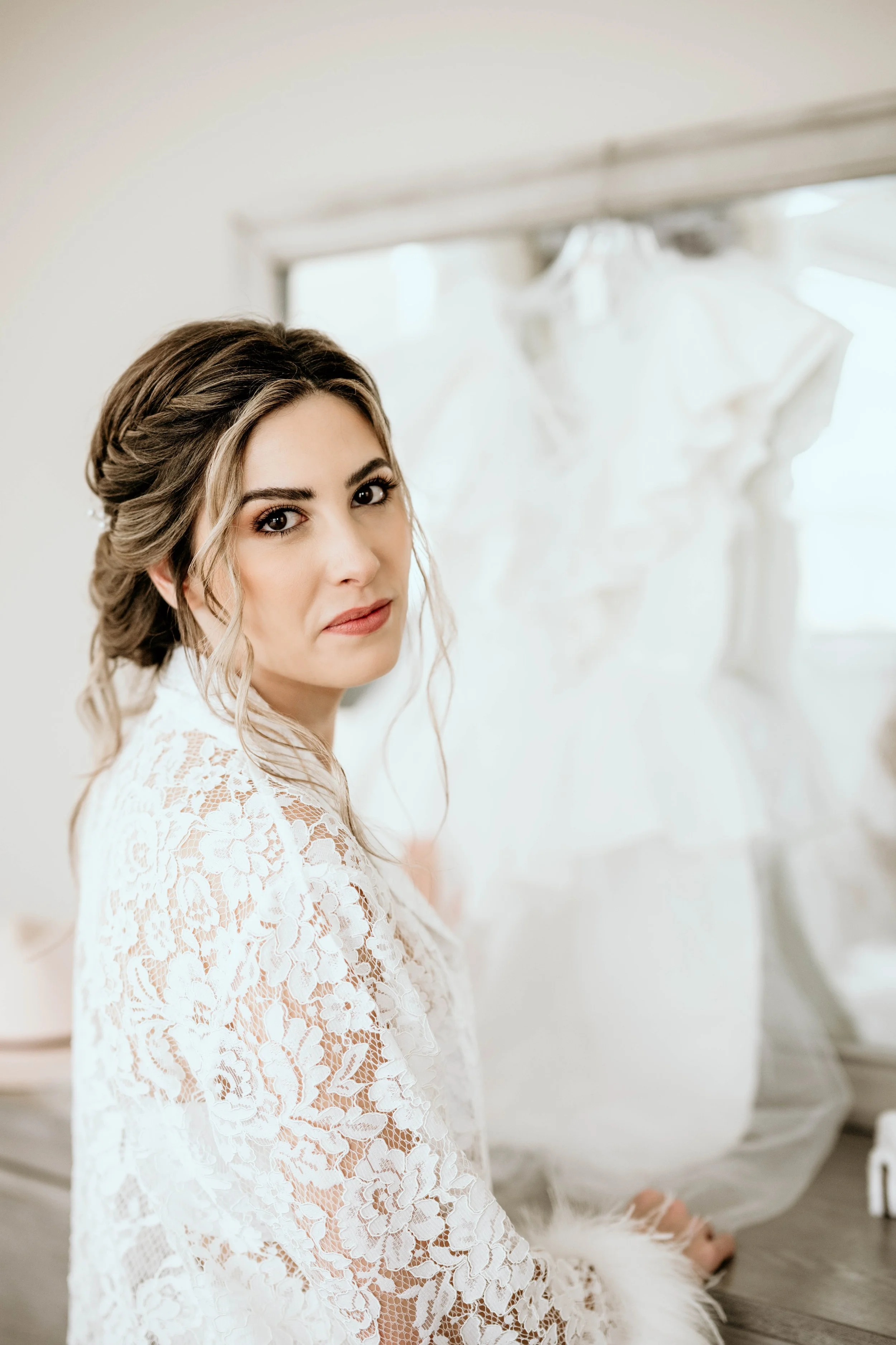 A woman in a white lace robe sits near a window with wedding dresses hanging in the background.Panama City beach weddings, PCB Photographers, PCB weddings