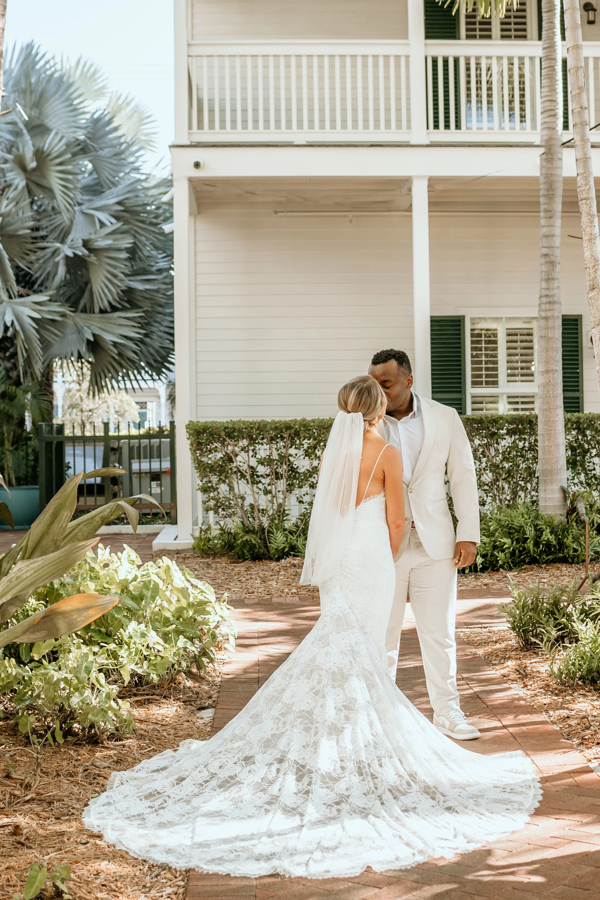 A bride in a white lace wedding dress and veil and a groom in a cream-colored suit kissing outdoors near a white house with green shutters.
