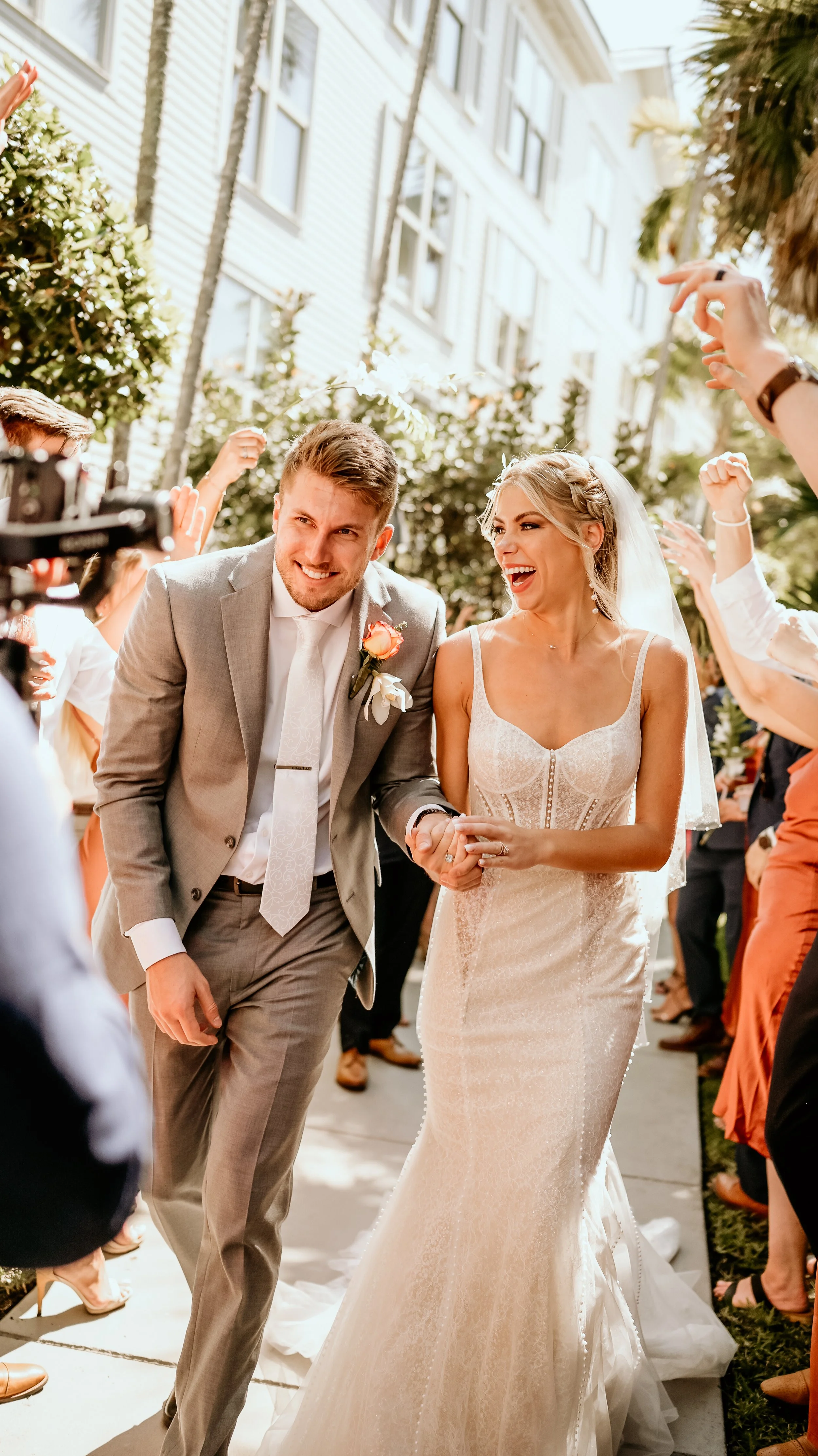 A newlywed couple walking hand in hand outdoors, smiling and celebrating, surrounded by friends and family.