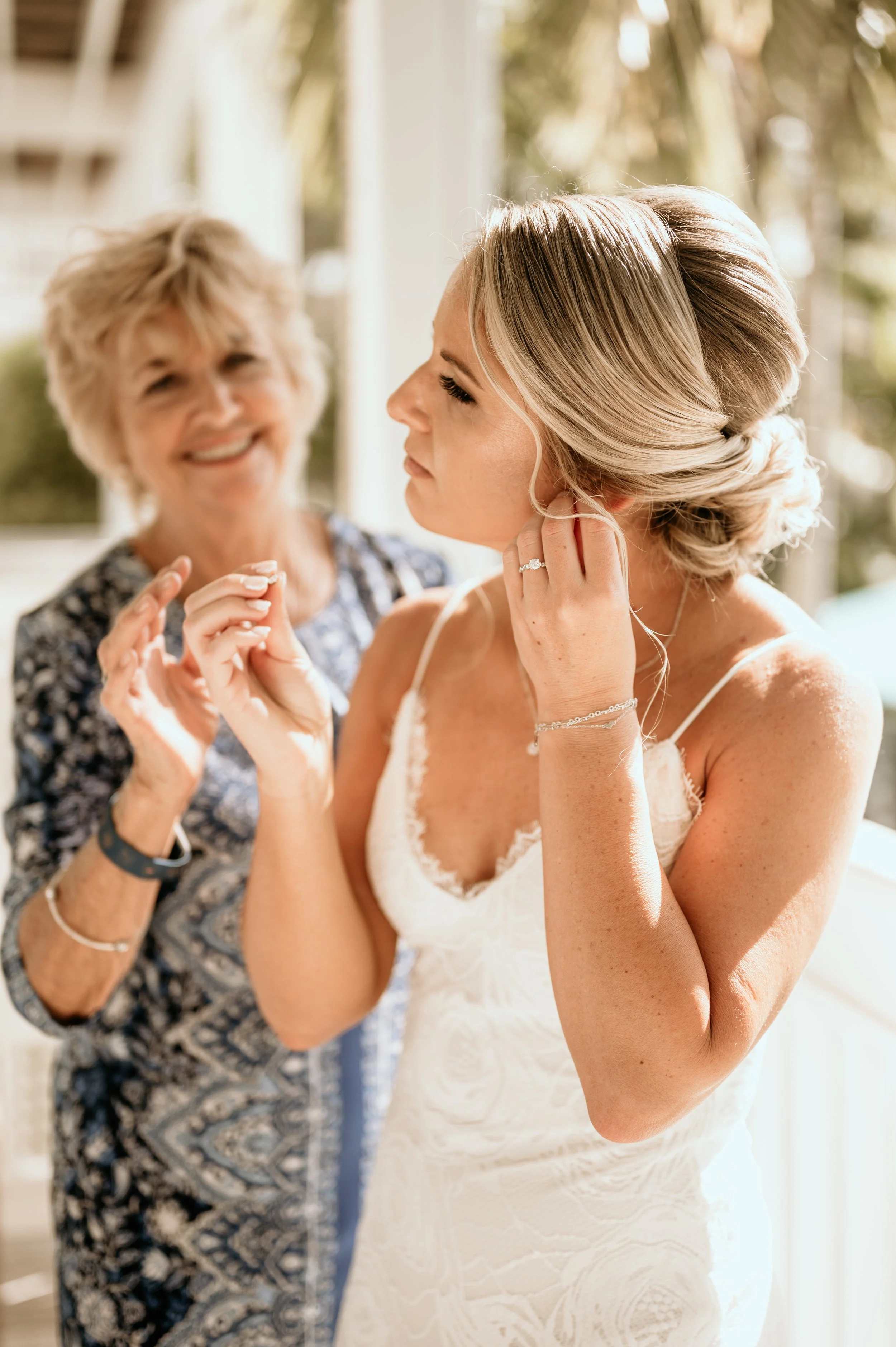 A bride in a white lace dress is getting ready, aided by an older woman, likely her mother or a close relative, on a porch with trees in the background.