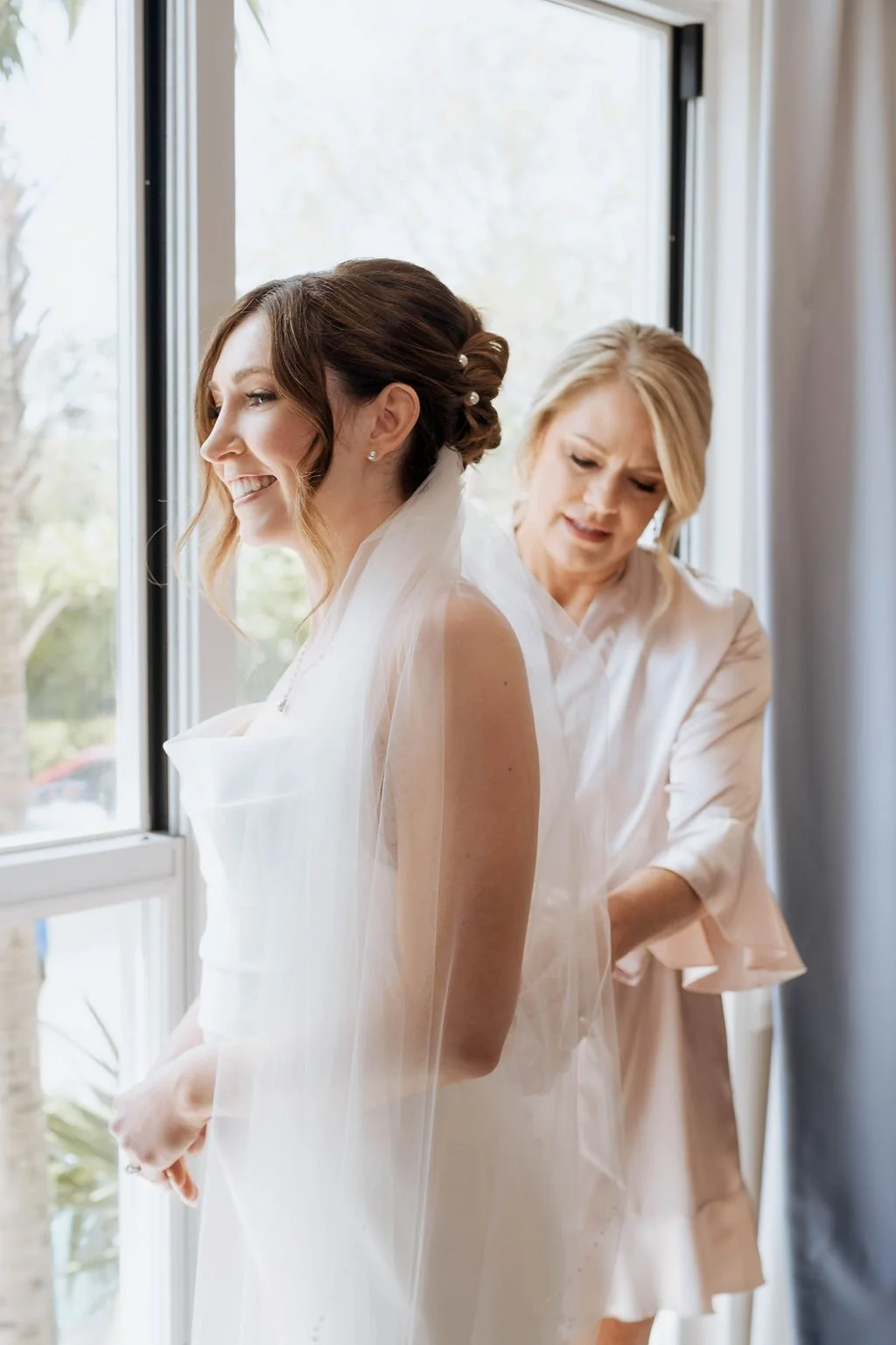 A bride smiling as she is assisted by a woman, possibly her mother, in a bedroom near a window, with greenery visible outside. St andrews state park weddings, Panama City beach weddings, PCB Photographers, PCB weddings
