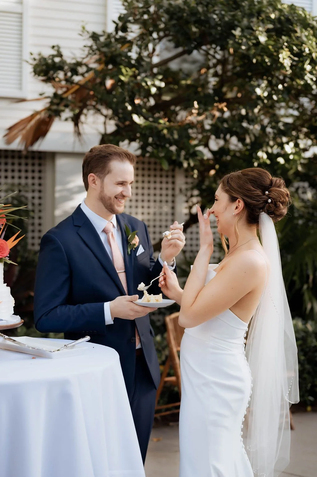 A bride and groom sharing a moment during their wedding cake celebration outdoors, with lush greenery and a white building in the background.
