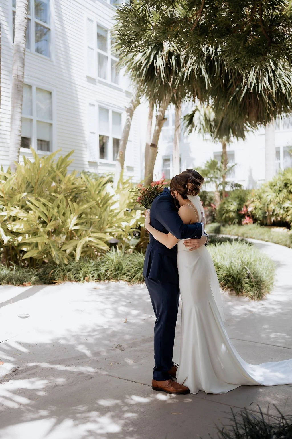 A bride and groom hugging outdoors in a garden area with lush green plants and palm trees.