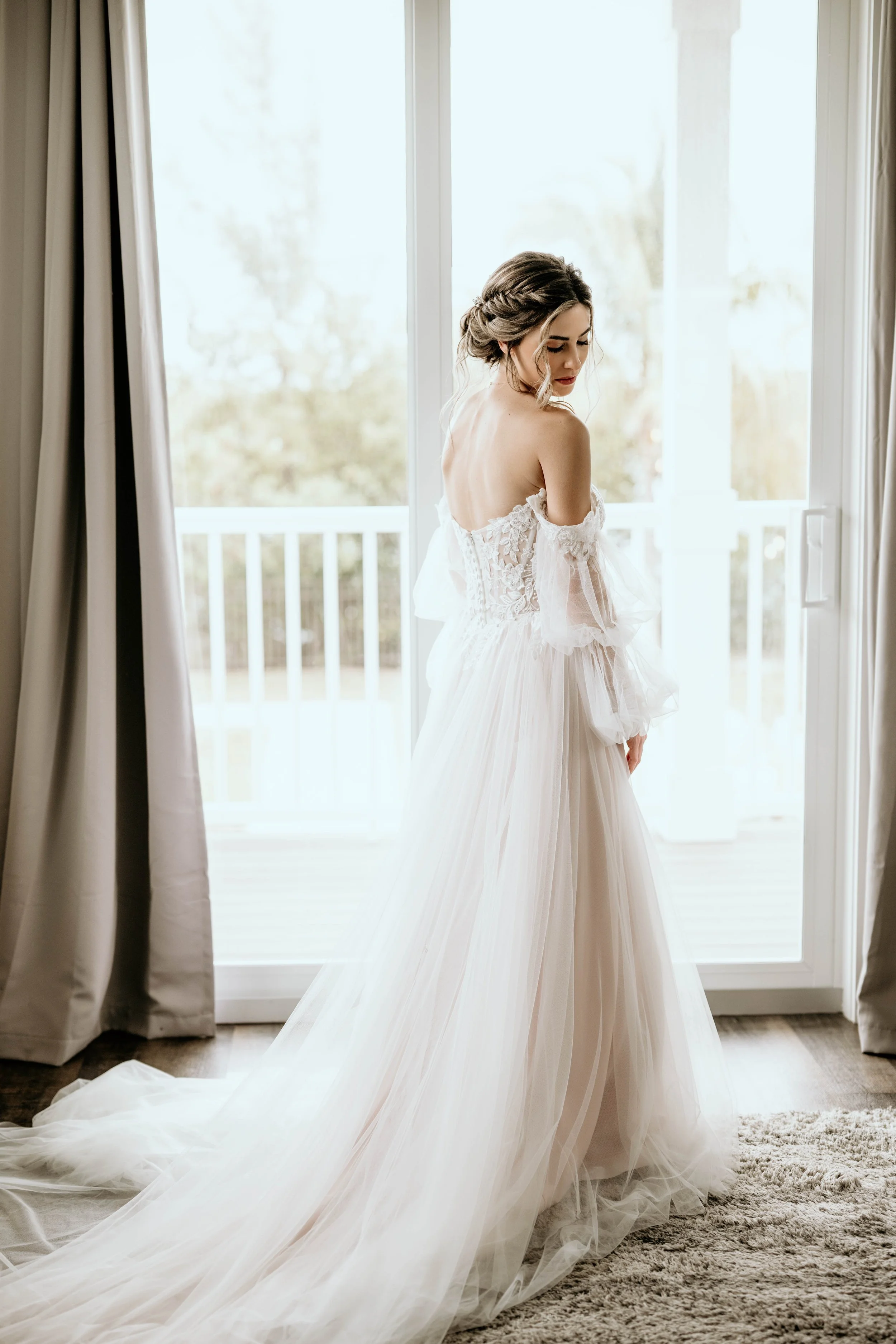 A bride in a white wedding gown standing in front of glass doors leading to a balcony, with curtains on either side and a plush carpet on the floor. St andrews state park weddings, Panama City beach weddings, PCB Photographers, PCB weddings
