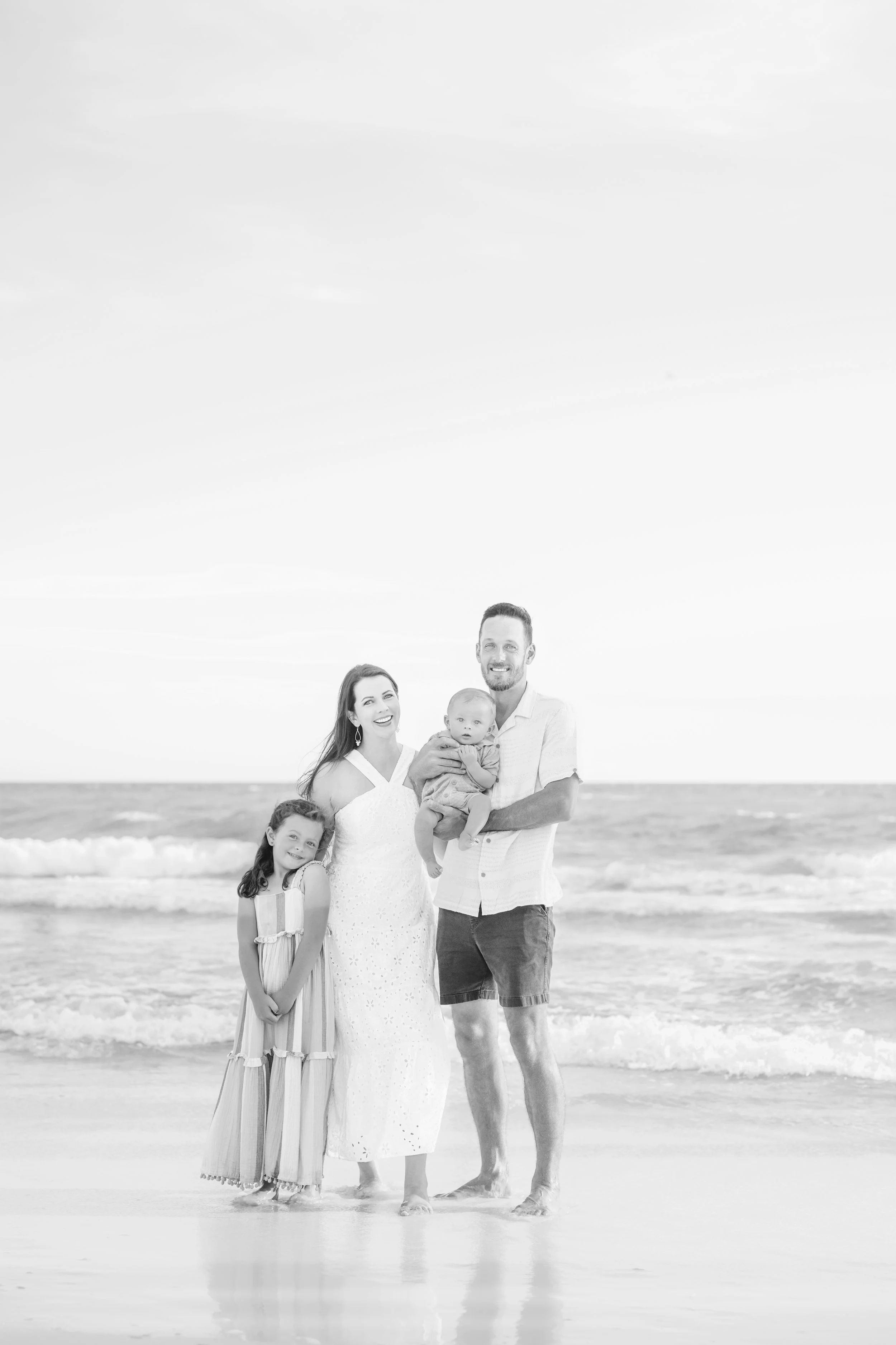 A smiling family of four standing on the beach in Panama City beach Florida for a photoshoot with ocean waves in the background, including a woman, a man holding a baby, and a young girl.