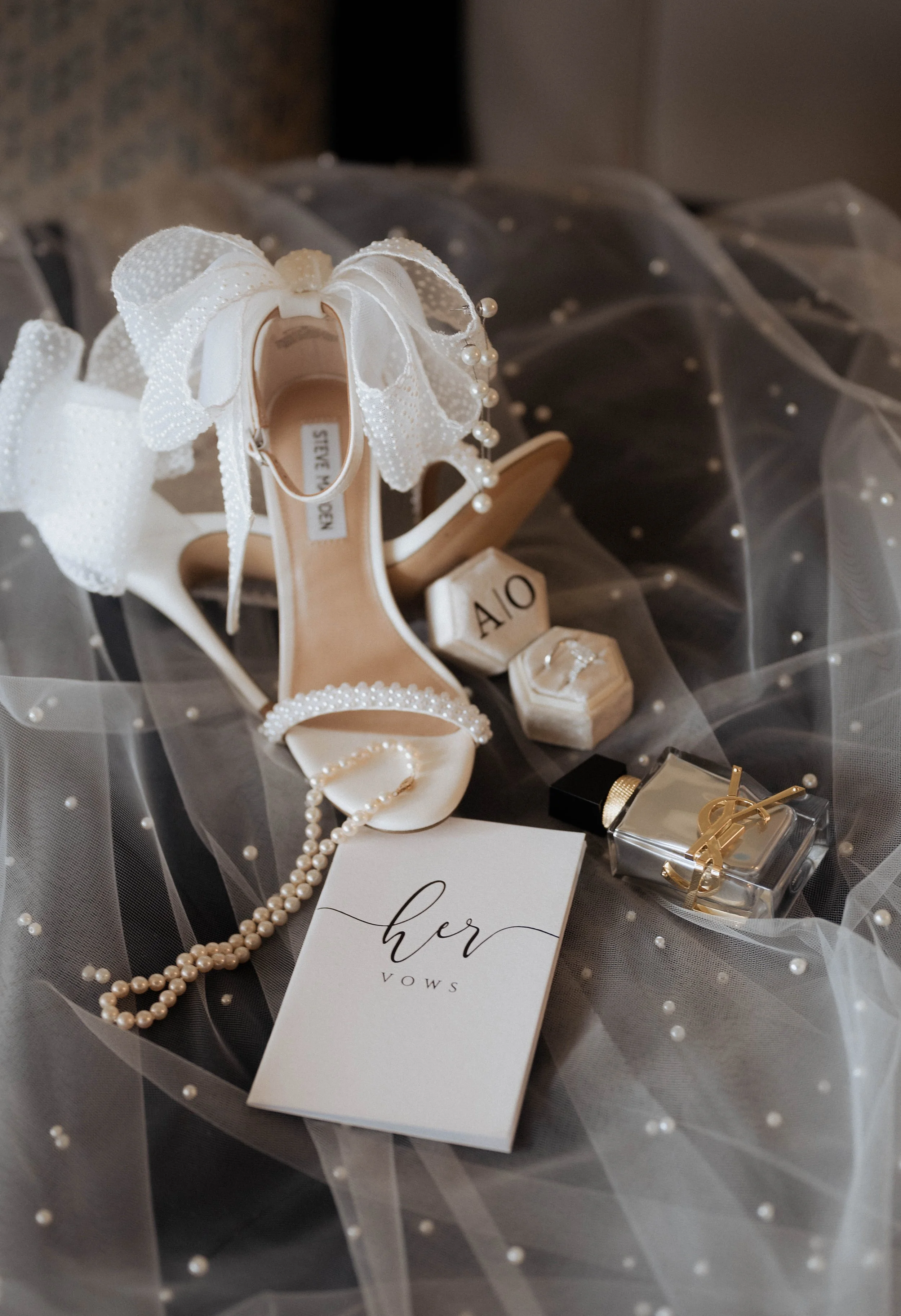 Wedding shoes decorated with bows and pearls, a vow card, pearl necklace, perfume, and jewelry on tulle fabric with pearl embellishments.