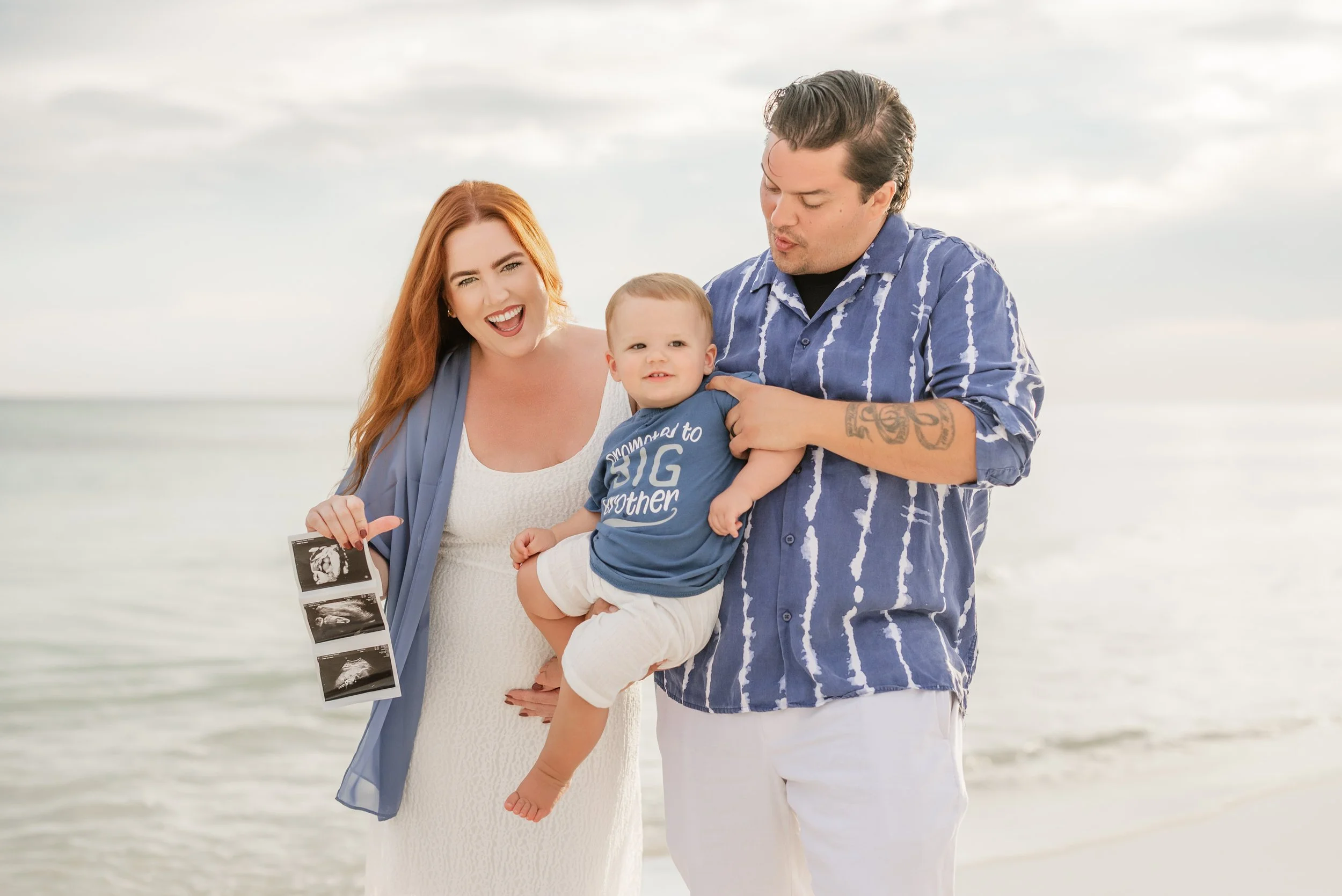 A happy family of three on the beach, holding ultrasound images, celebrating a pregnancy in Panama City beach florida. The mother has red hair, the father has dark hair and tattoos.