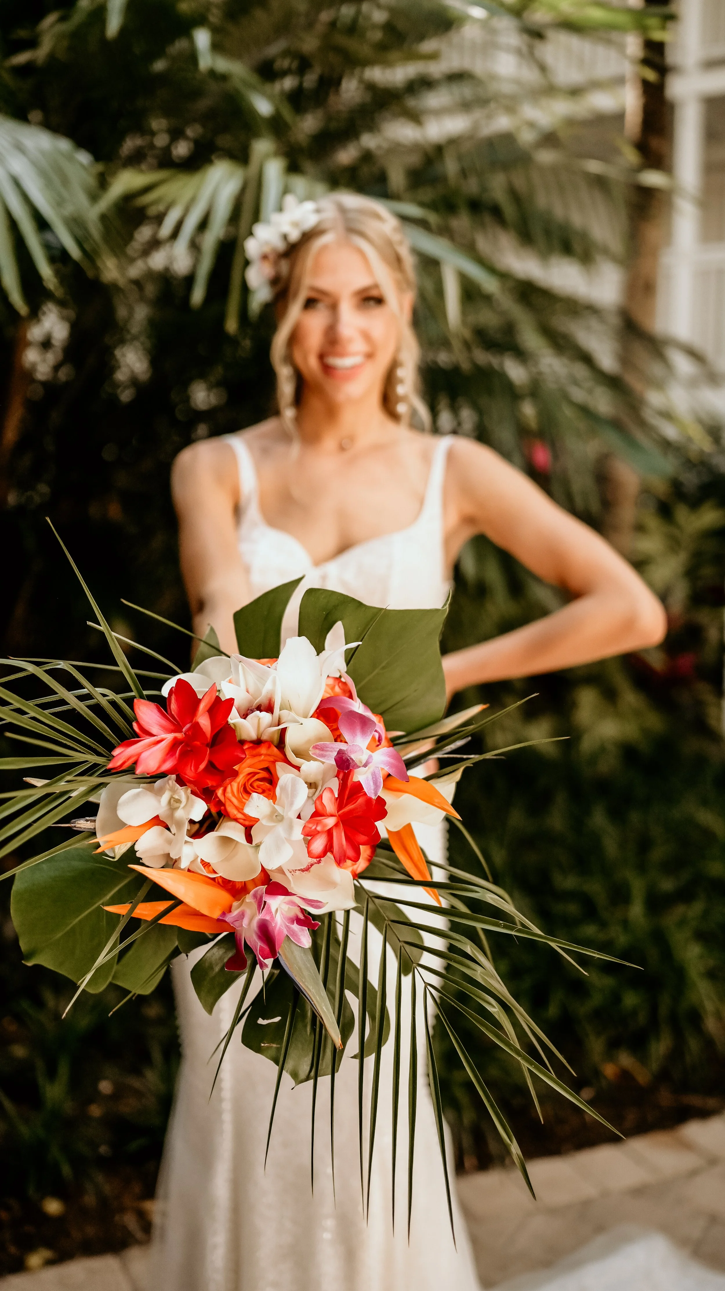 A smiling bride in a white wedding dress holding a large tropical bouquet with flowers and greenery, standing outdoors with lush green foliage in the background.
