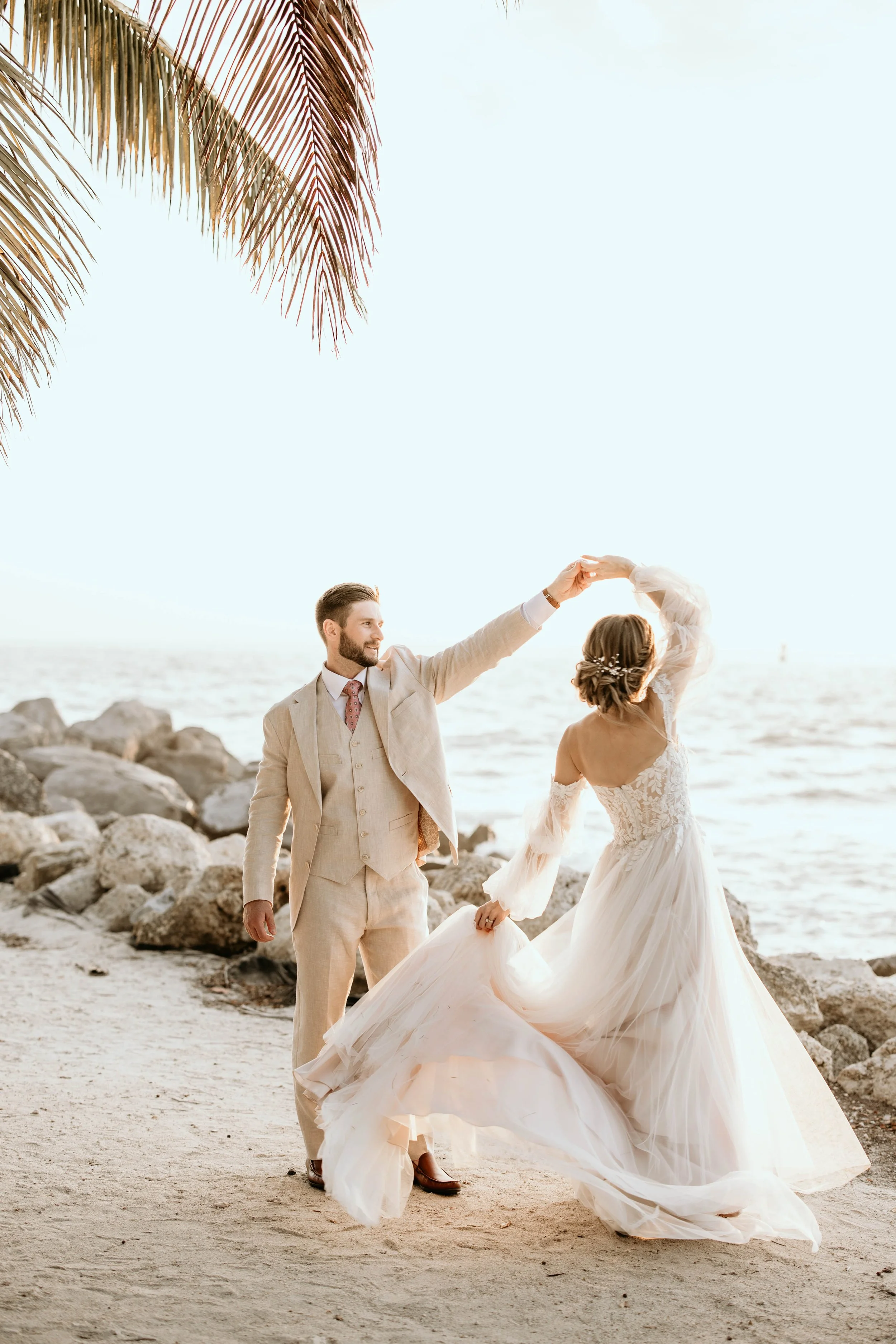 A couple dancing on a beach at sunset, with the man wearing a beige suit and the woman in a flowing white wedding gown. Panama City beach wedding photographer