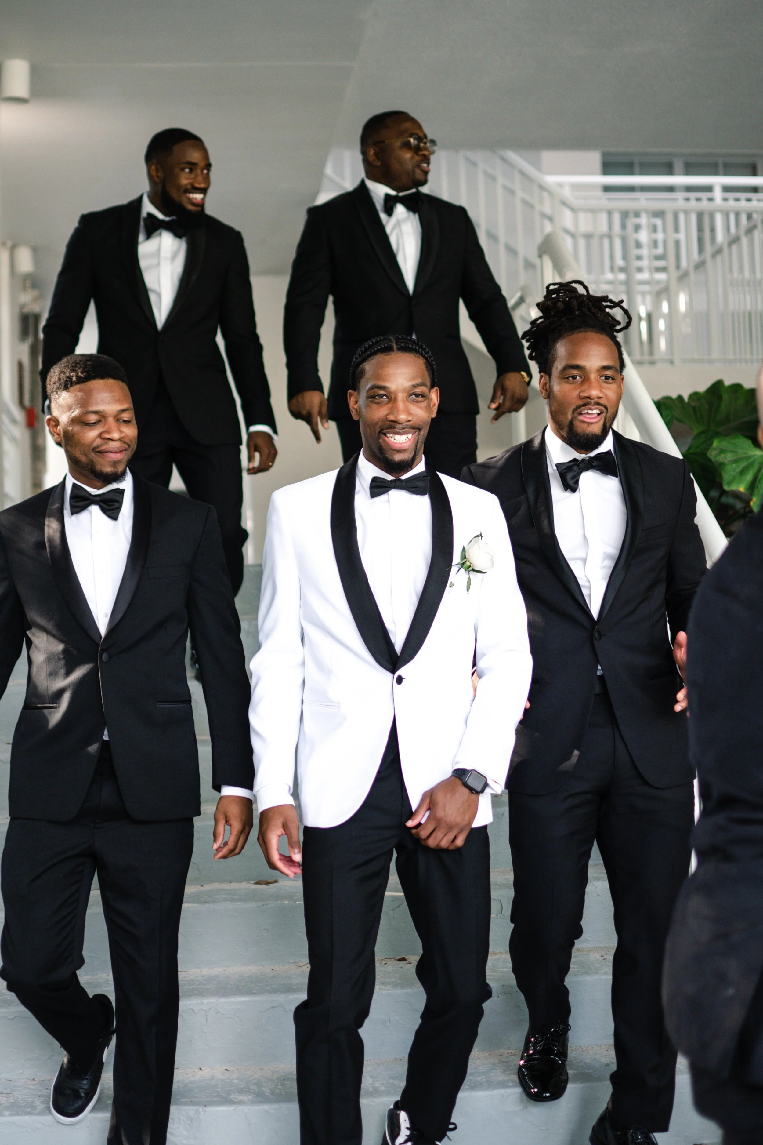 Group of men dressed in tuxedos, some with bow ties, celebrating and smiling on a staircase in a modern building.