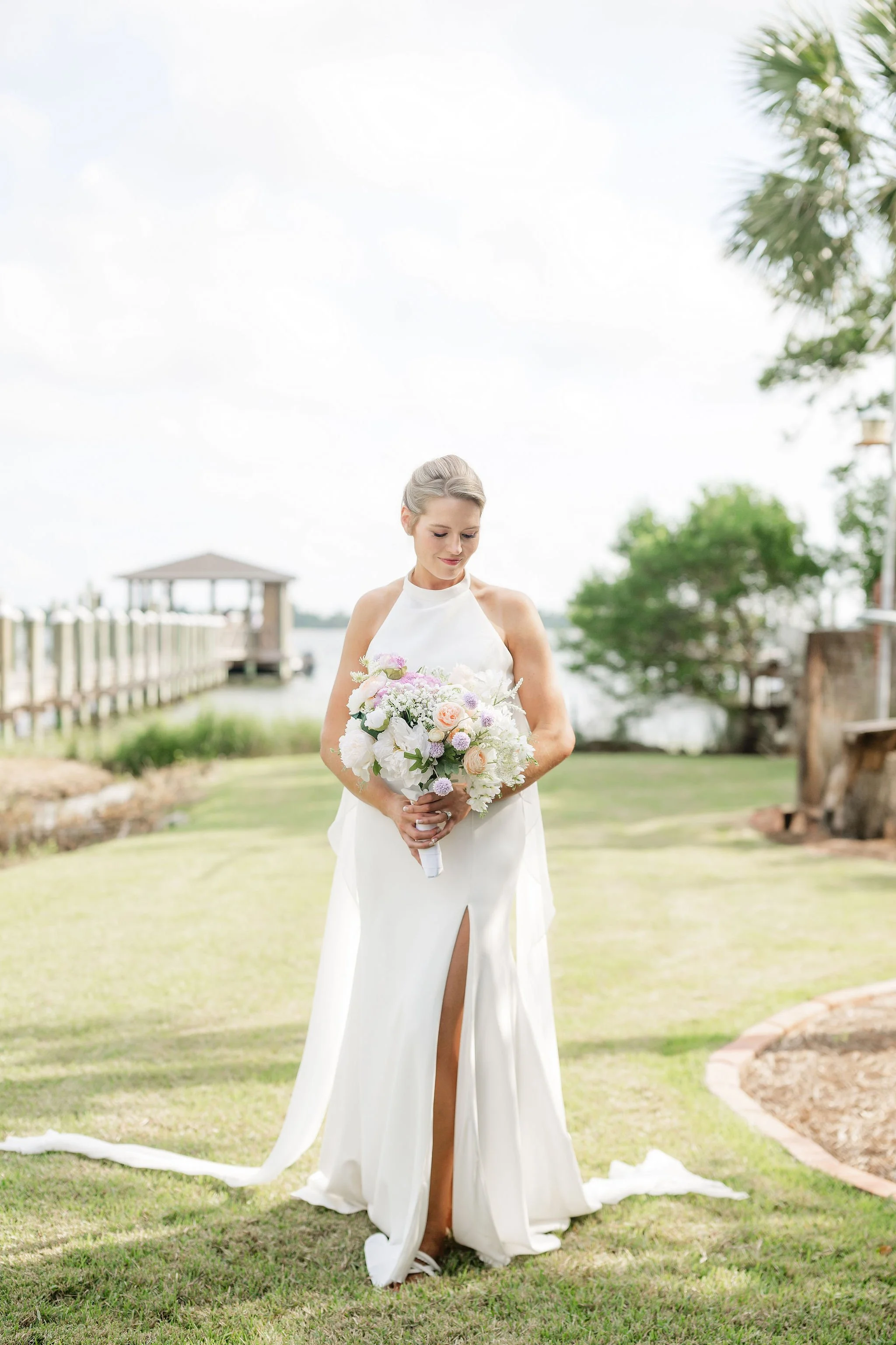 A bride standing outdoors in a white wedding gown holding a bouquet of flowers with a slitted front. She is on a grassy area near a body of water with trees and a wooden gazebo in the background. Panama City beach wedding photographer