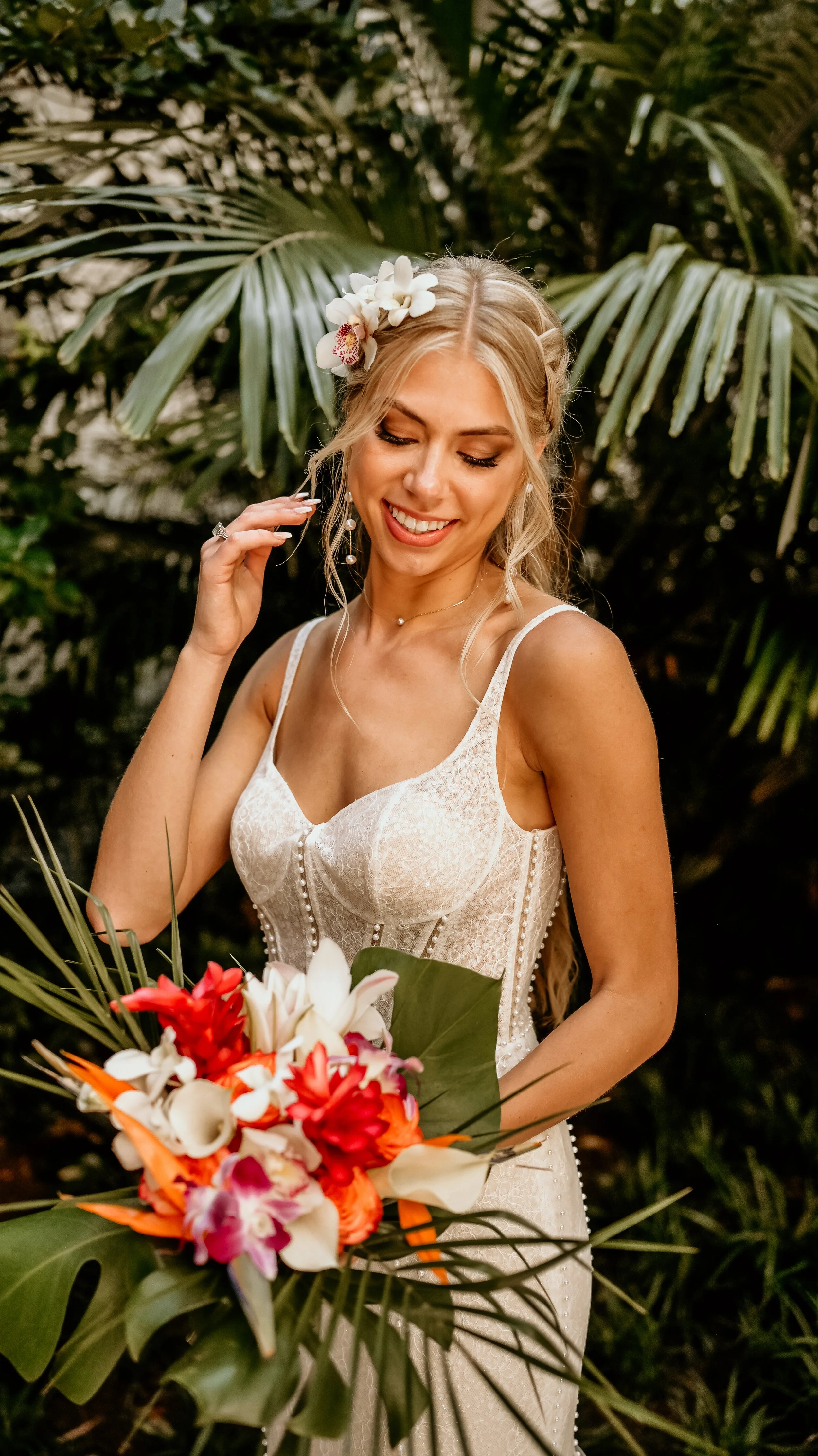 A bride holding a colorful tropical bouquet, standing in front of lush green foliage, smiling with her eyes closed.