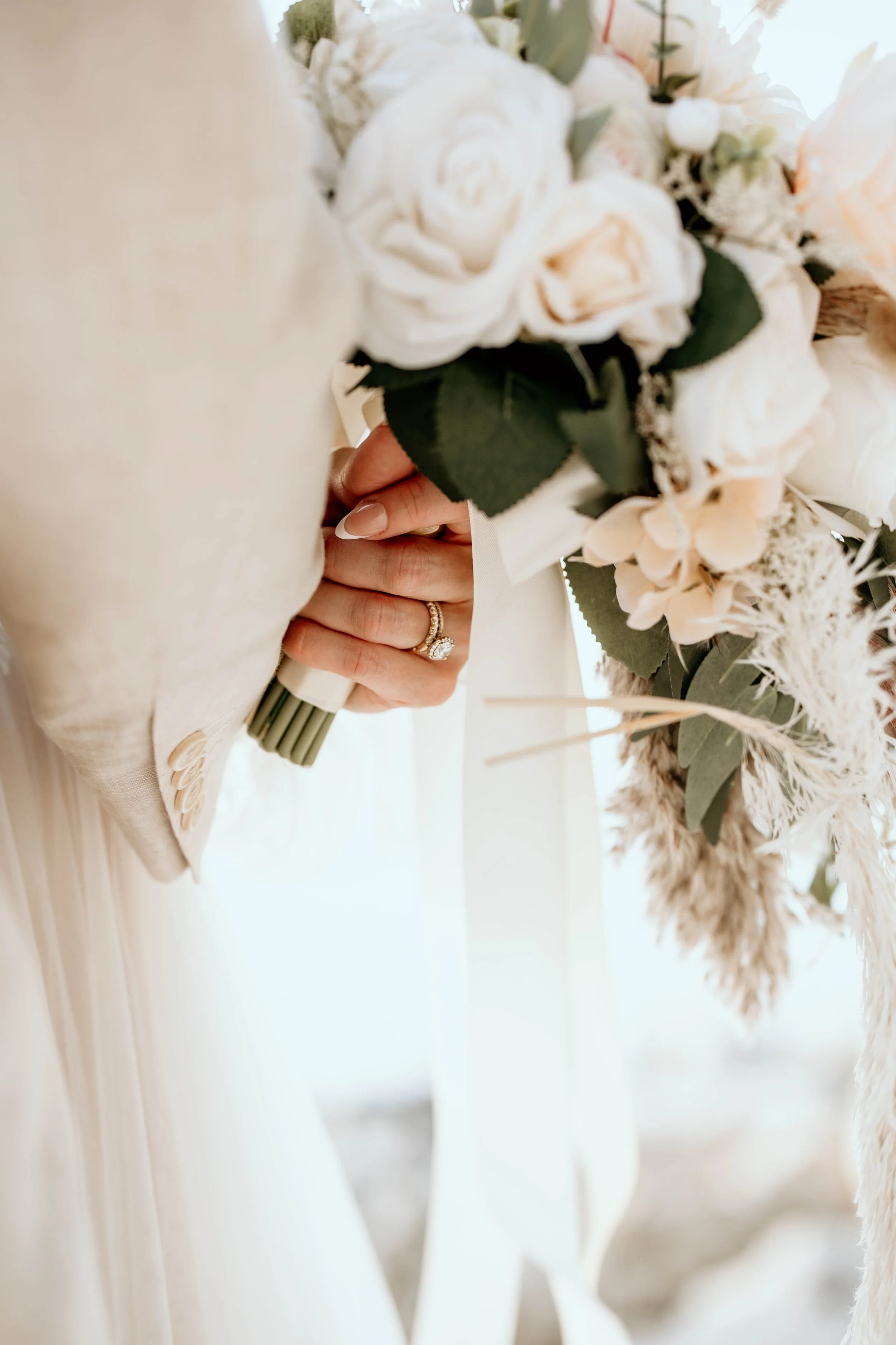 Close-up of a woman holding a large bouquet of white and pale pink roses and greenery, with multiple rings on her fingers visible. Panama City beach wedding