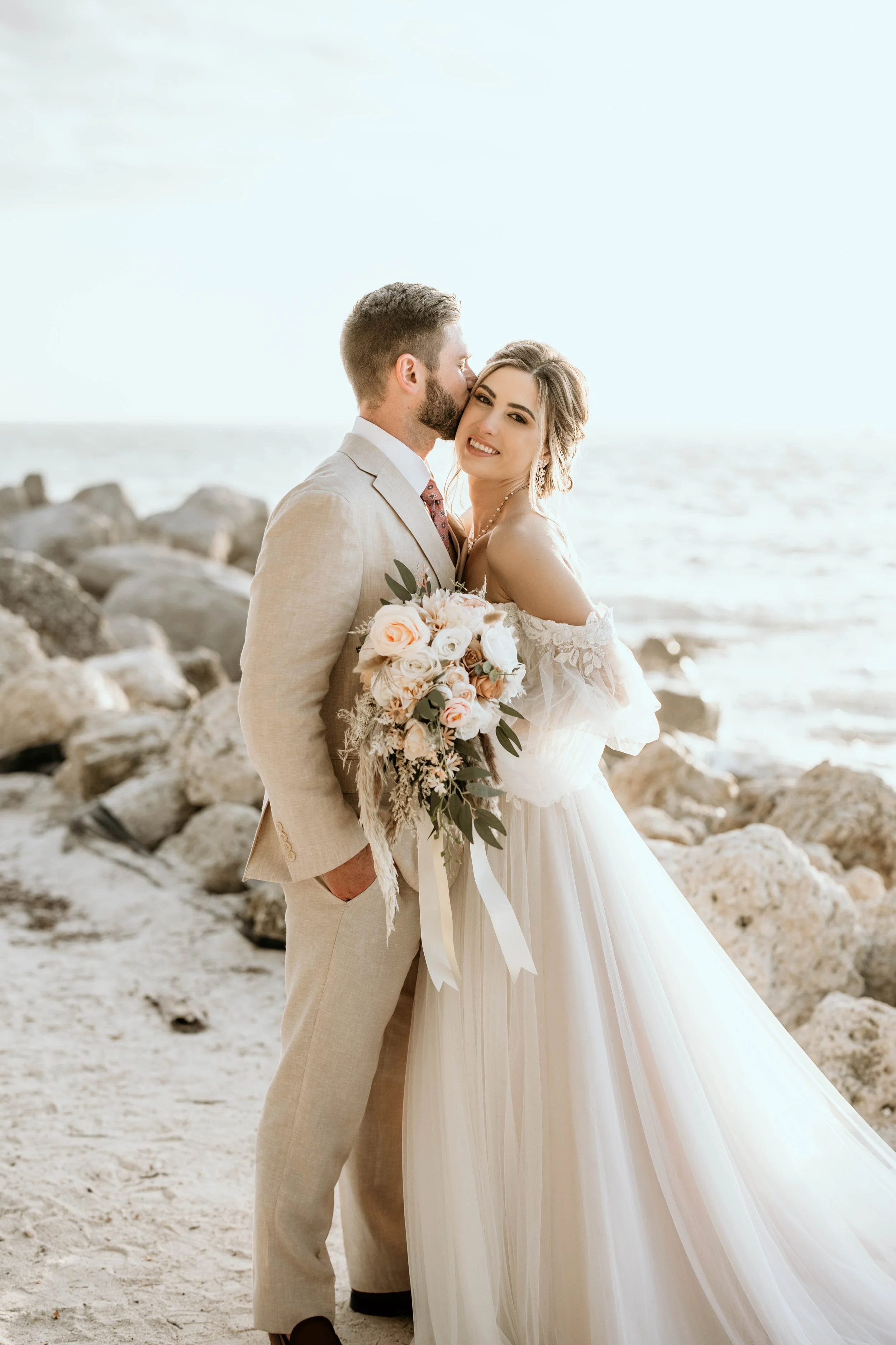 A newlywed couple poses in Panama City beach florida The groom is whispering in the bride's ear while she holds a bouquet of flowers. The bride is smiling and wearing a flowing white wedding gown, and the groom is in a light suit. panam