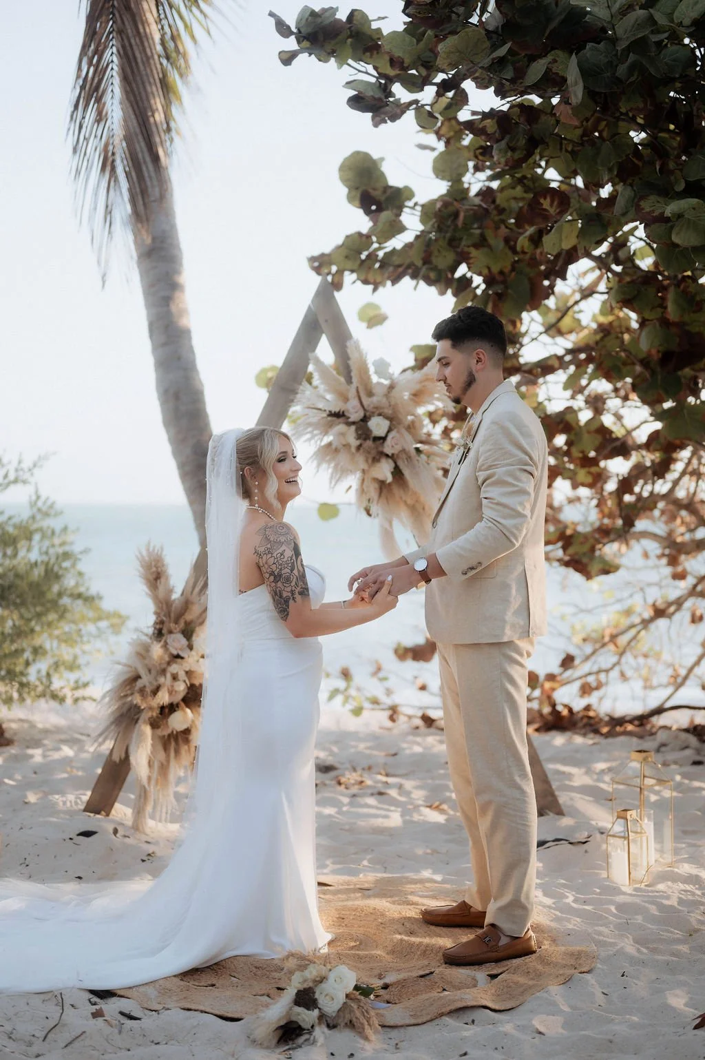 A couple getting married on a beach, holding hands and exchanging vows, with a wooden wedding arch decorated with pampas grass and flowers behind them. The bride is wearing a white wedding gown and veil, and the groom is in a beige suit. The scene is