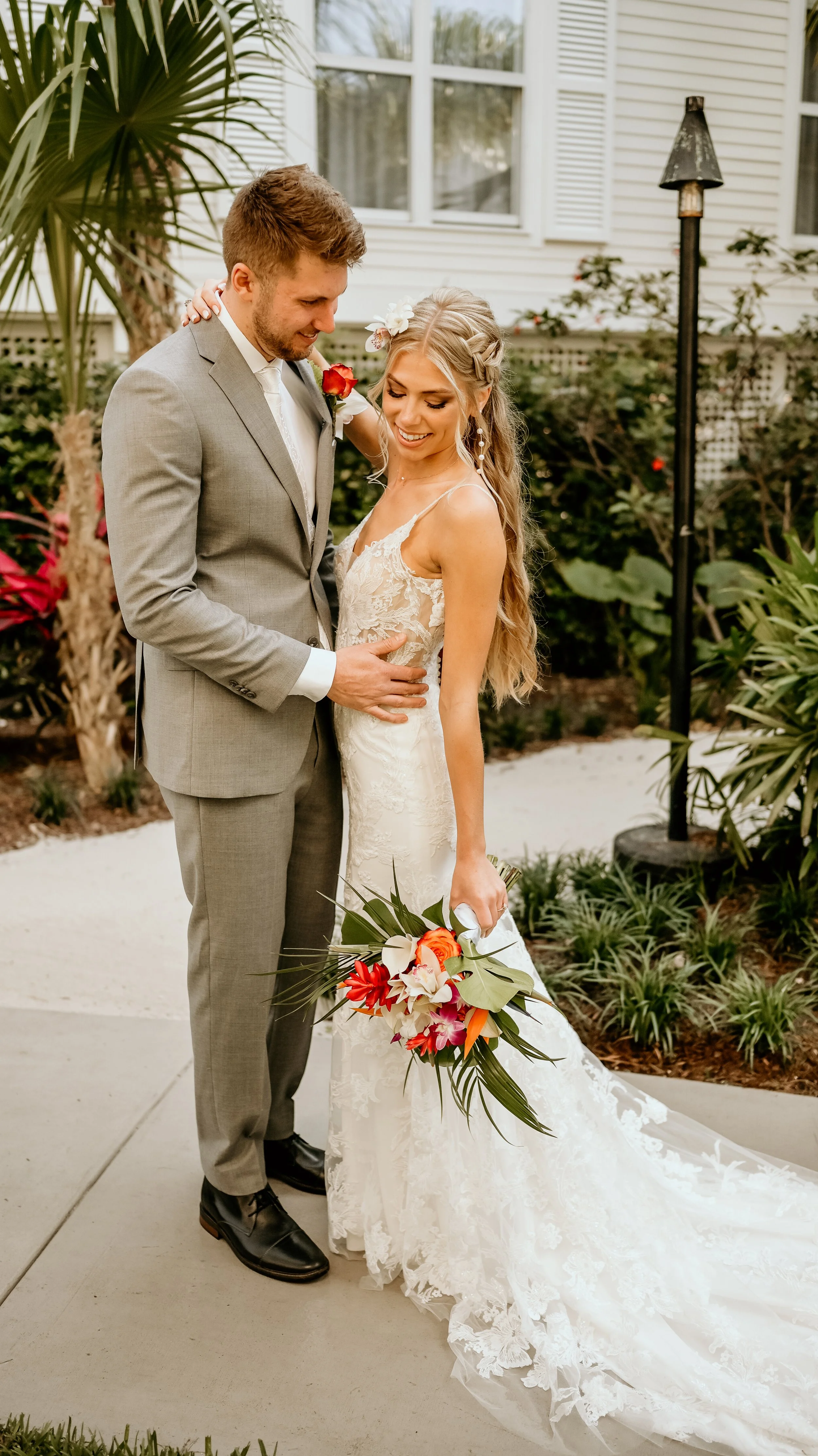 A bride and groom in wedding attire sharing a tender moment outdoors, with the bride holding a bouquet of tropical flowers.