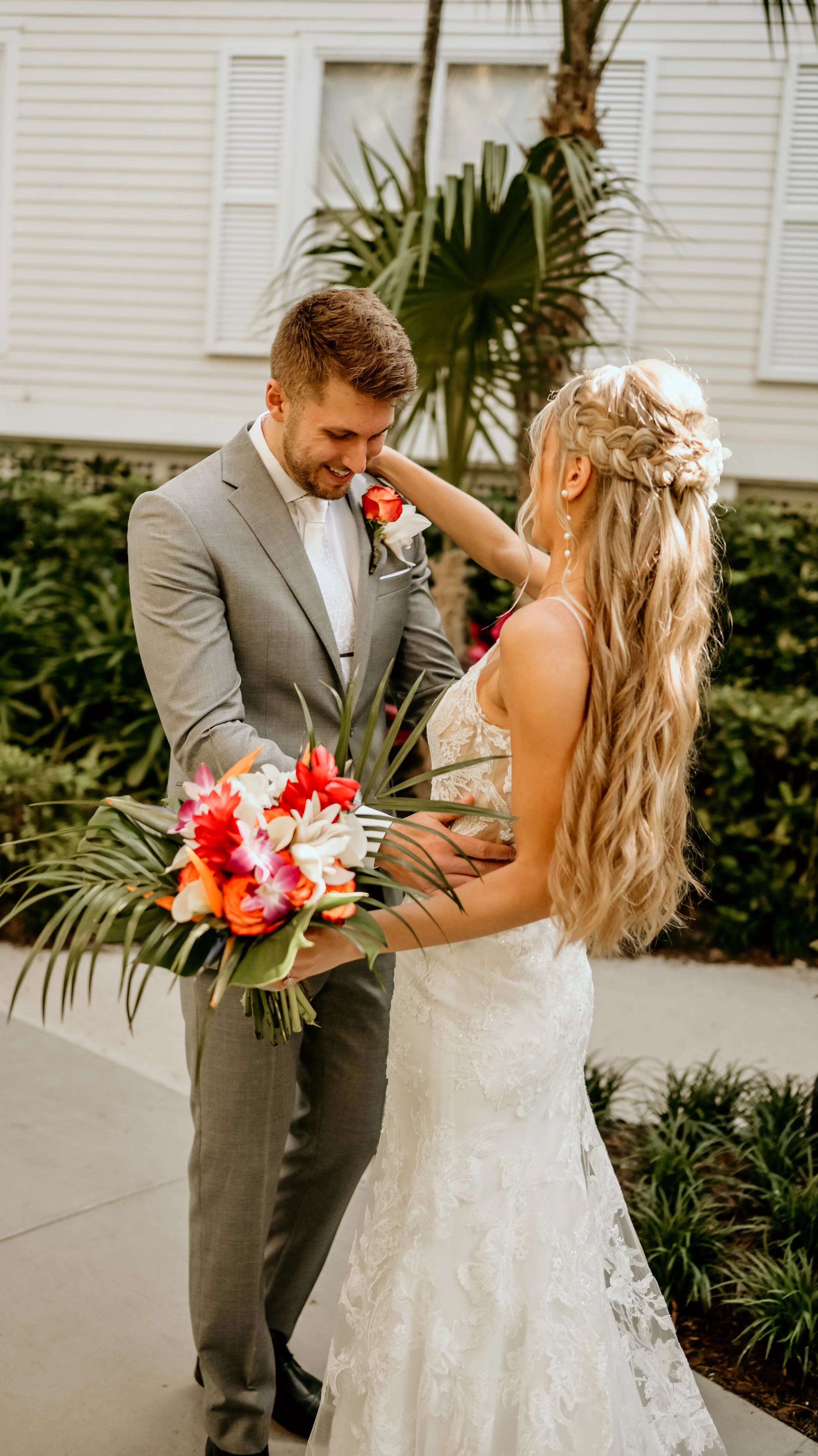 A bride and groom during their wedding, with the groom smiling and holding a vibrant bouquet and the bride touching his face gently, outdoors near a house with greenery.