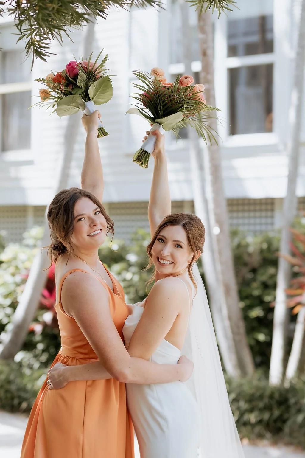 Two women celebrating, one in a white dress, and the other in a peach dress, holding bouquets above their heads. weddings, Panama City beach weddings, PCB Photographers, PCB weddings