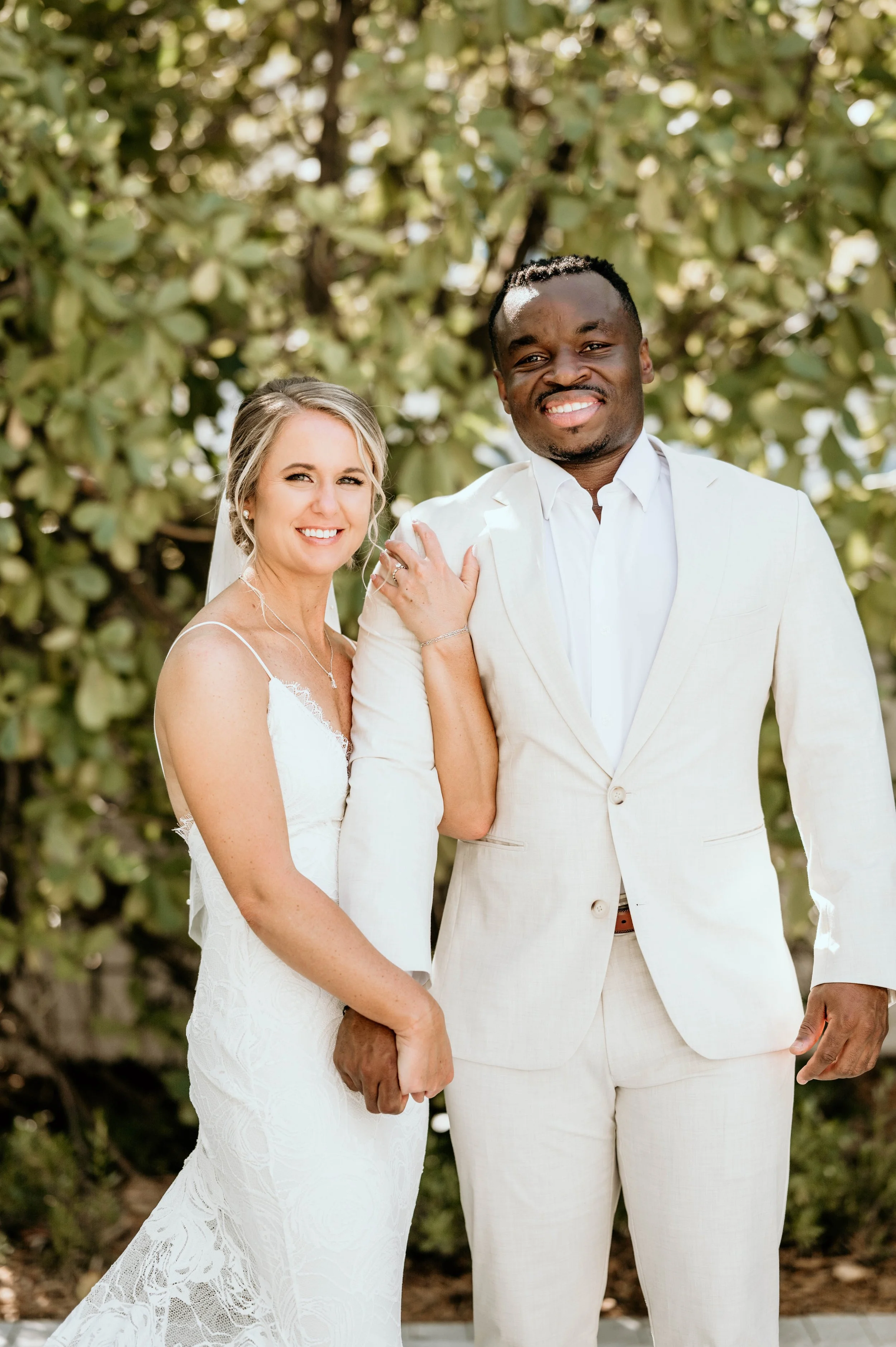 A smiling newlywed couple stands outdoors in front of green foliage. The bride wears a white lace wedding dress, and the groom is dressed in a light-colored suit with a white shirt.