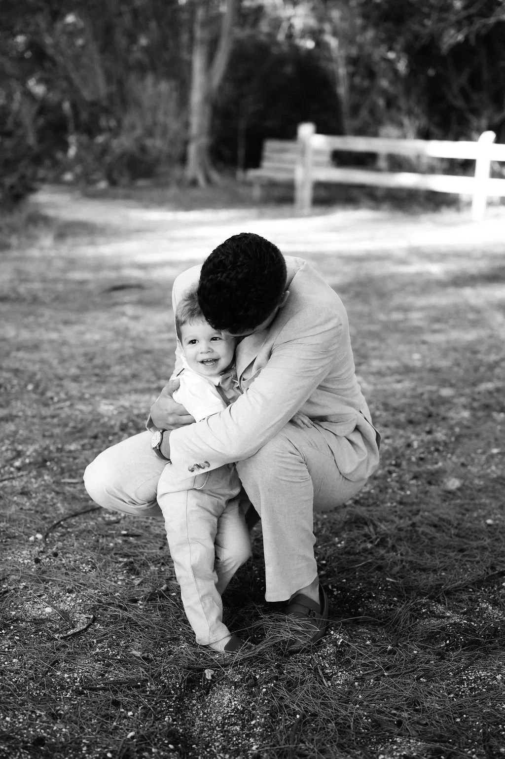 A man is crouched down, hugging a smiling young boy in an outdoor setting with trees and a wooden fence in the background.