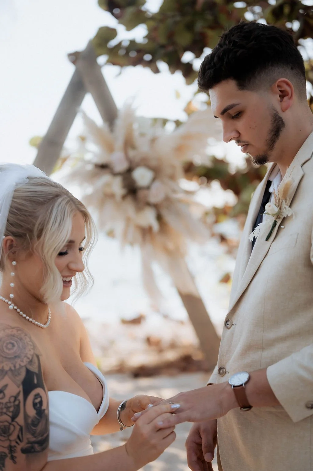 A bride and groom exchange rings during a wedding ceremony on a beach, with a wooden arch decorated with beige and white flowers in the background.