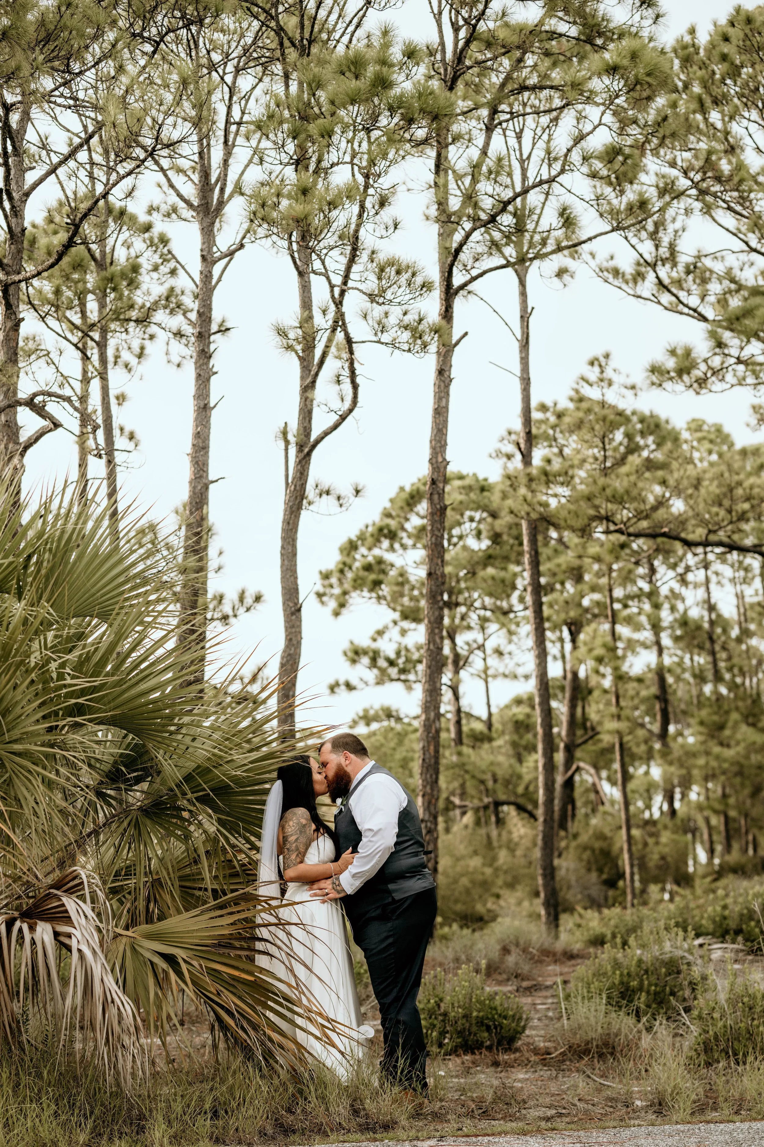 A couple sharing a kiss in a wooded outdoor setting on their wedding day, with tall pine trees and bushes in the background. St andrews state park weddings, Panama City beach weddings, PCB Photographers, PCB weddings