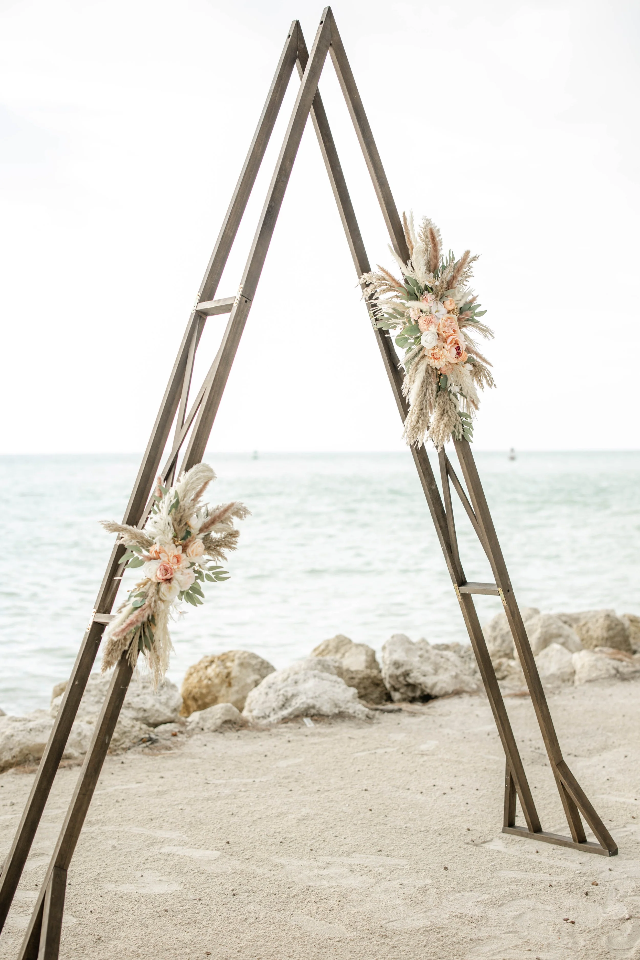 Wooden wedding arch decorated with pink and white flowers and pampas grass on a sandy beach with rocks and the ocean in the background. St andrews state park weddings, Panama City beach weddings, PCB Photographers, PCB weddings