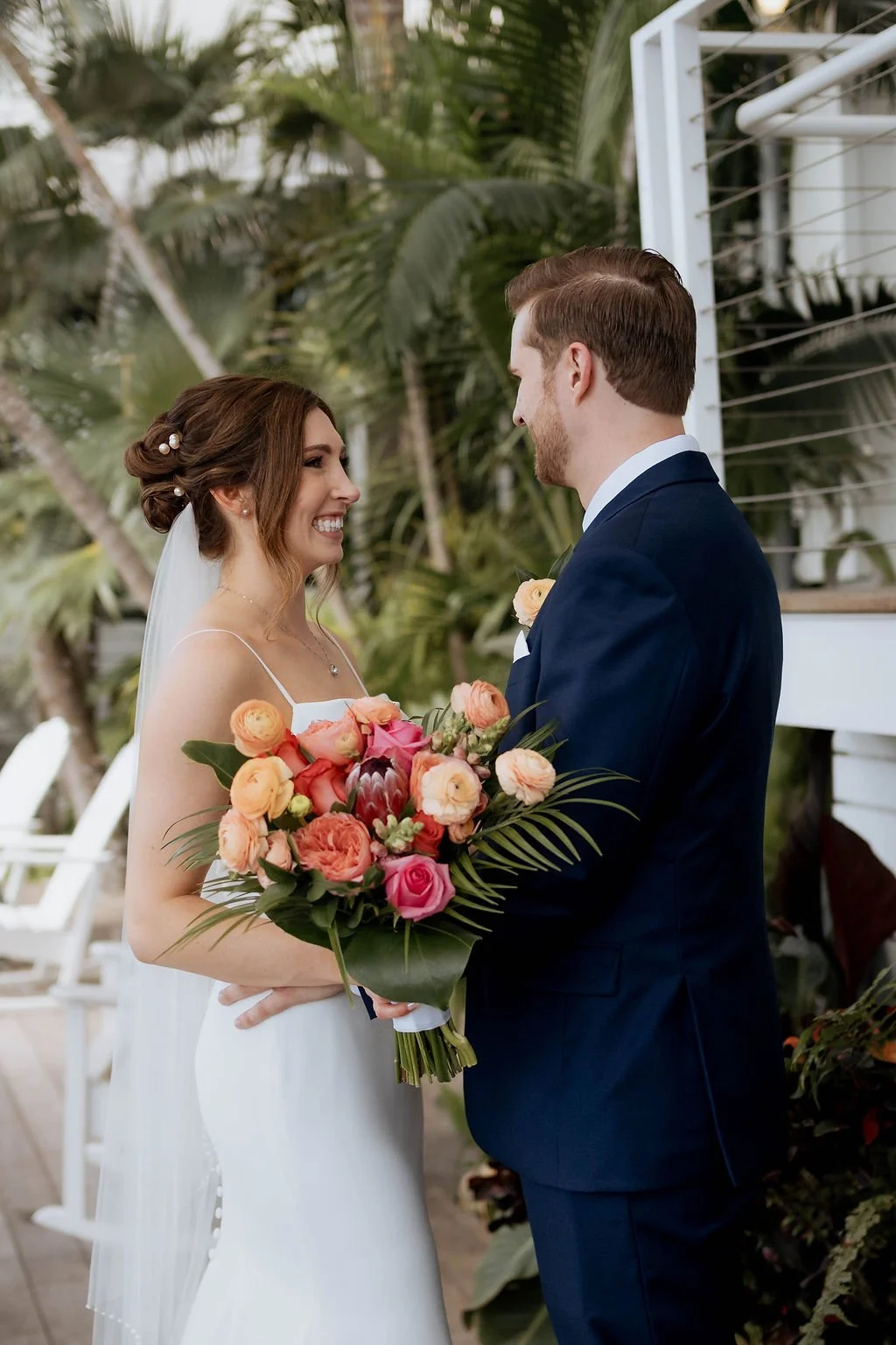 A bride in a white wedding dress and a groom in a navy suit exchange vows outdoors, with the bride holding a large bouquet of pink, peach, and coral flowers, surrounded by lush greenery.
