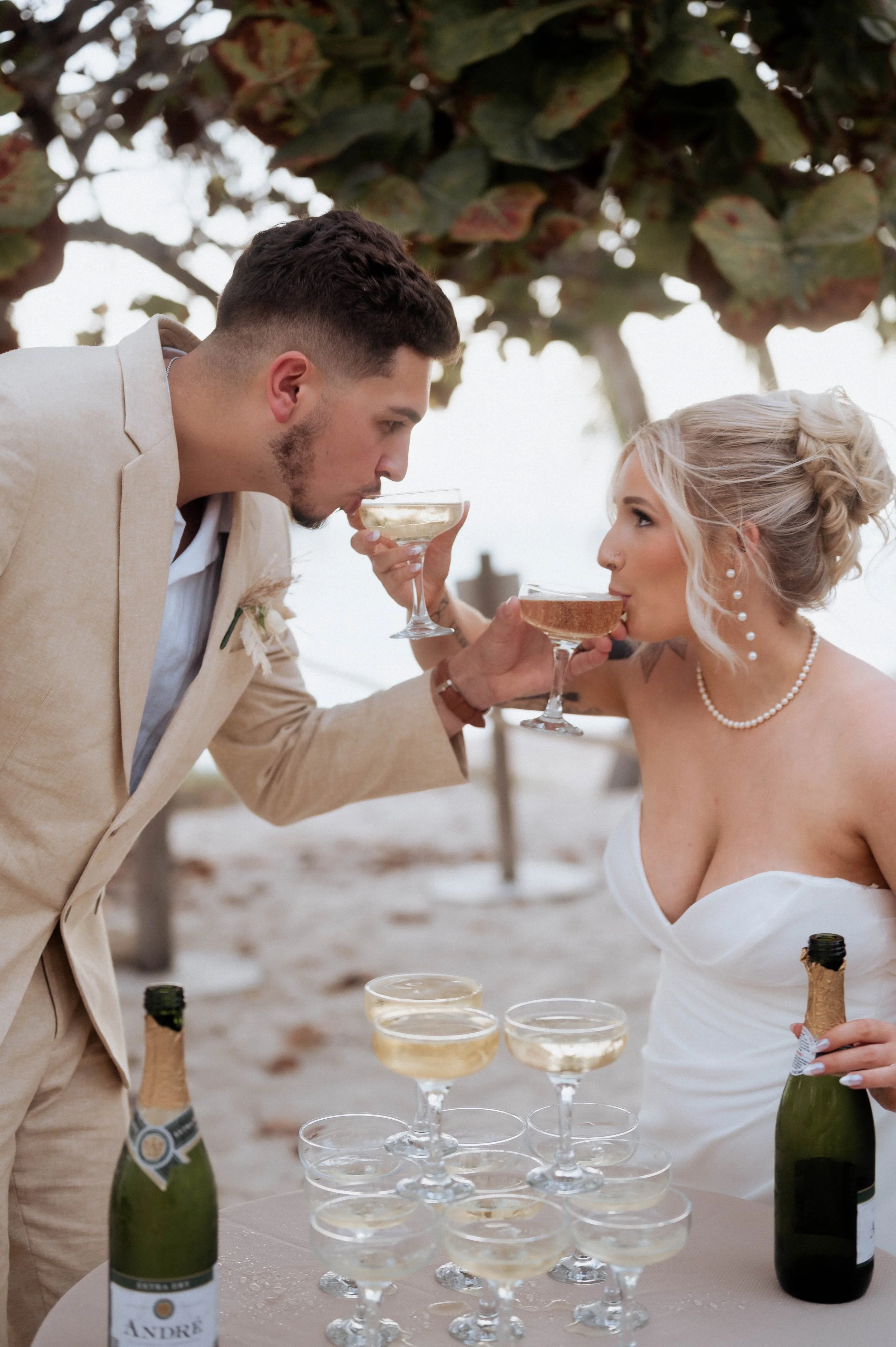 A bride and groom at their beach wedding, sharing a toast with champagne. They are holding coupe glasses, and there are bottles of champagne on the table with additional glasses ready for the toast.