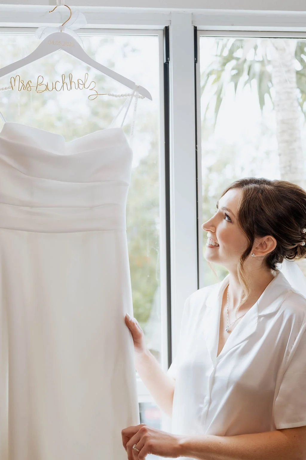 A woman with brown hair and a white shirt looking at a white wedding dress on a hanger with a personalized 'Mrs. Buchholz' in gold wire. St andrews state park weddings, Panama City beach weddings, PCB Photographers, PCB weddings
