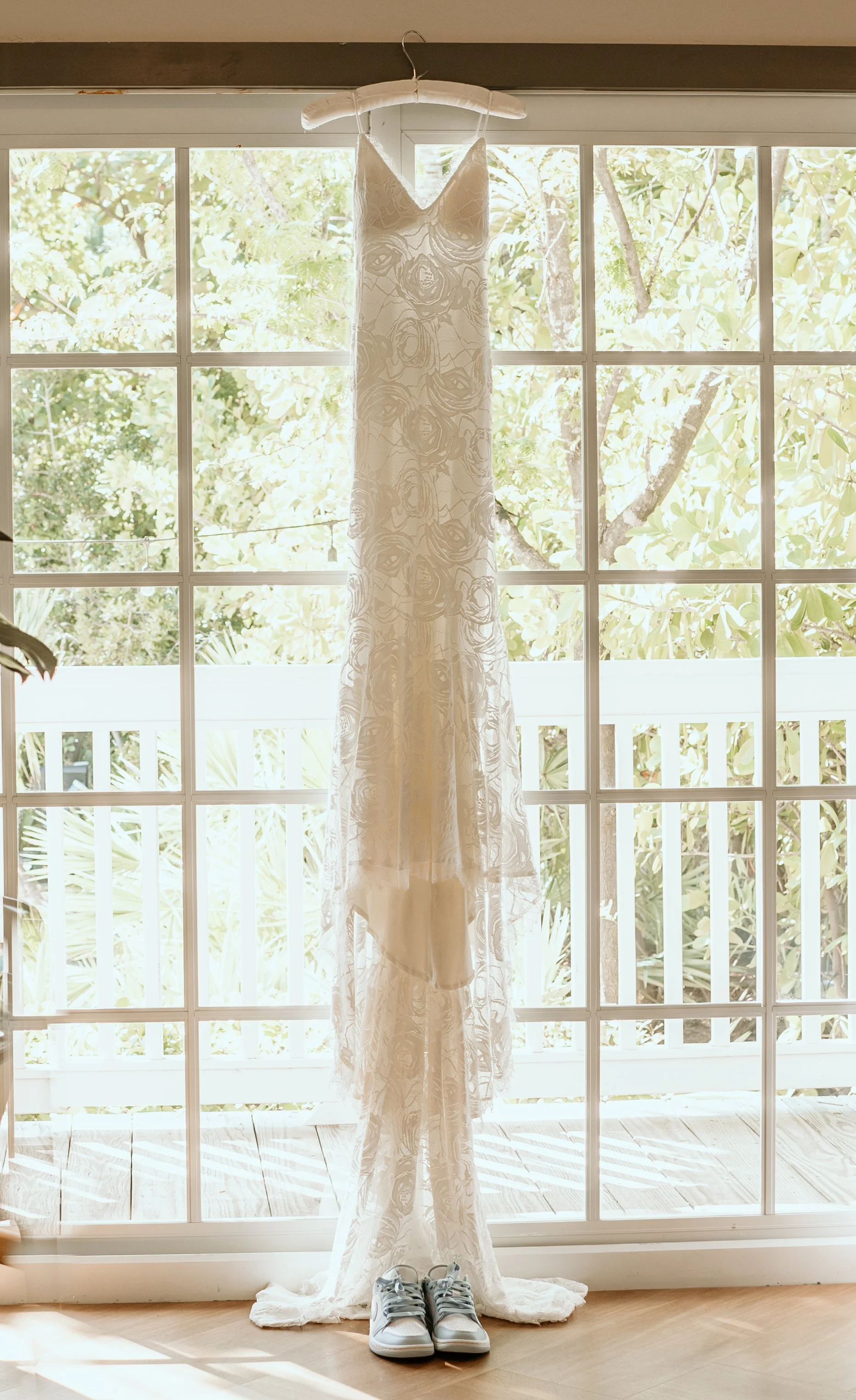 A wedding dress hanging on a hanger, with white sneakers placed underneath it in front of a large window with a view of trees and greenery outside.