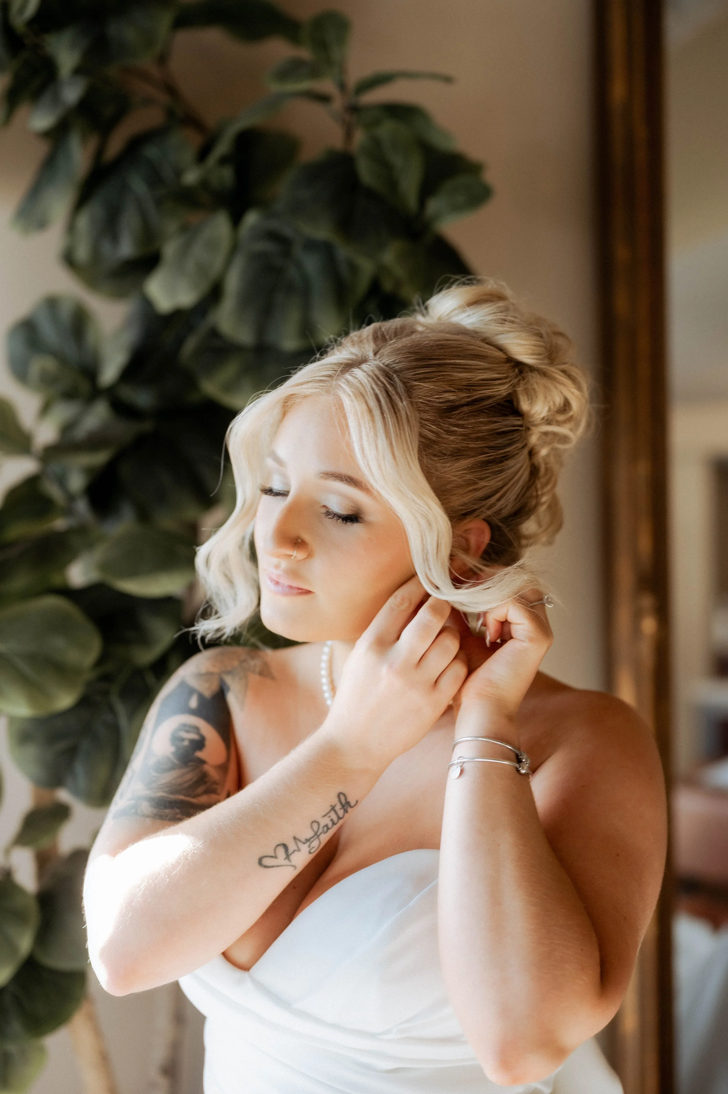 A woman with blonde hair in loose waves, wearing a white strapless dress, adjusting her earring in front of a large leafy green plant, with sunlight illuminating her face and shoulder.