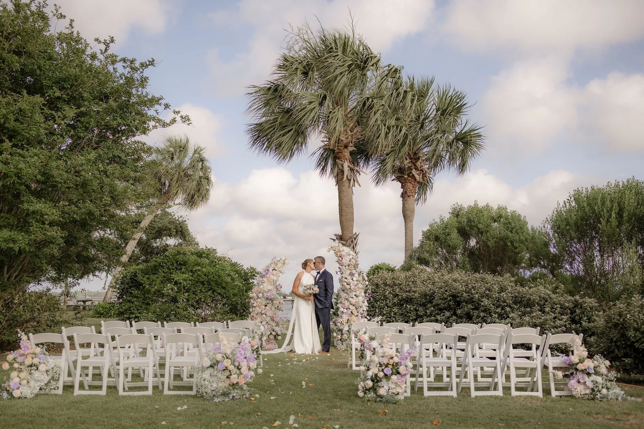 A wedding ceremony outdoors with a couple standing at the altar, surrounded by flowers, palm trees, and chairs for guests. floral from coastal bloom rentals