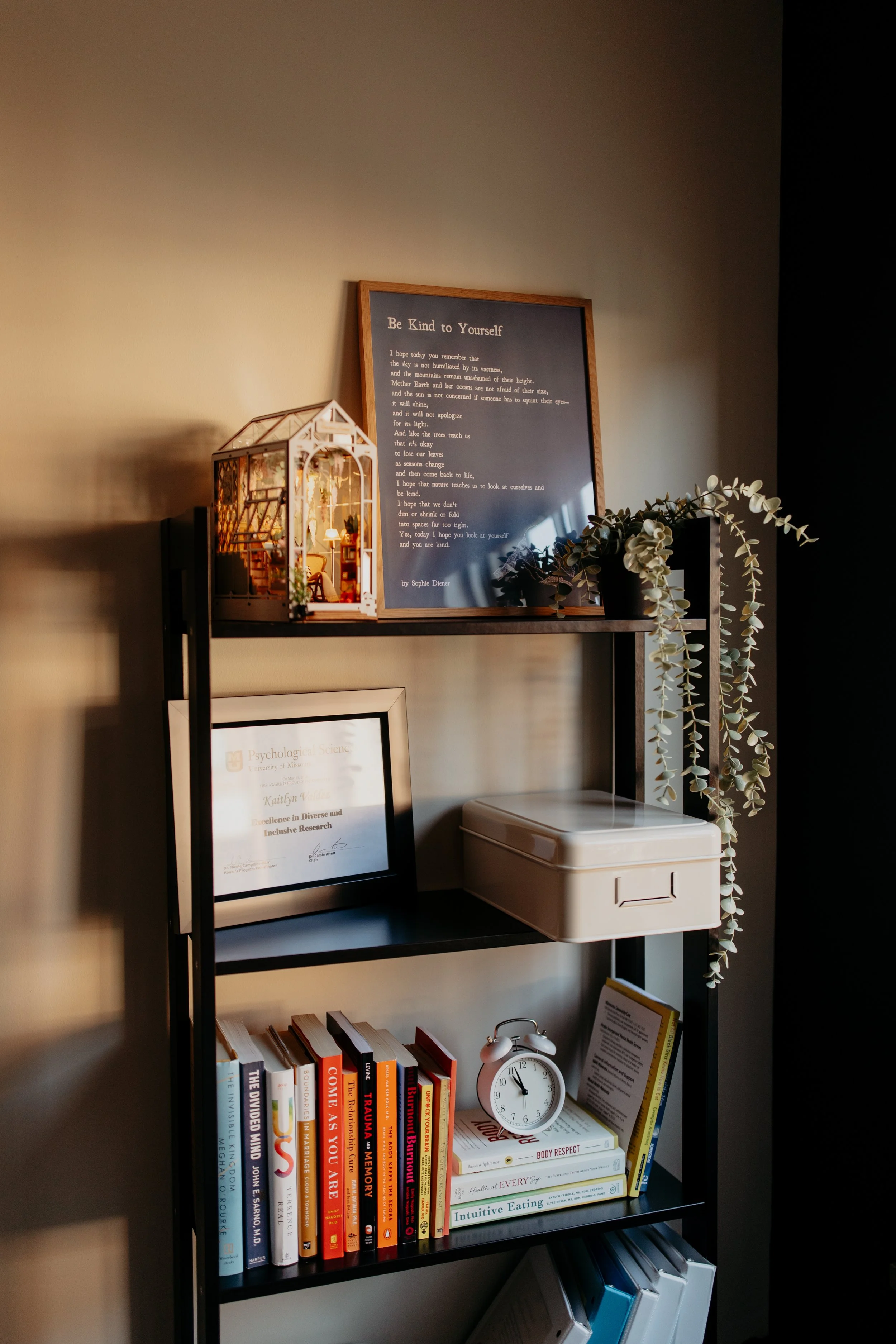 Interior shelf with framed certificate, books, clock, miniature house, poem, and decorative plant.