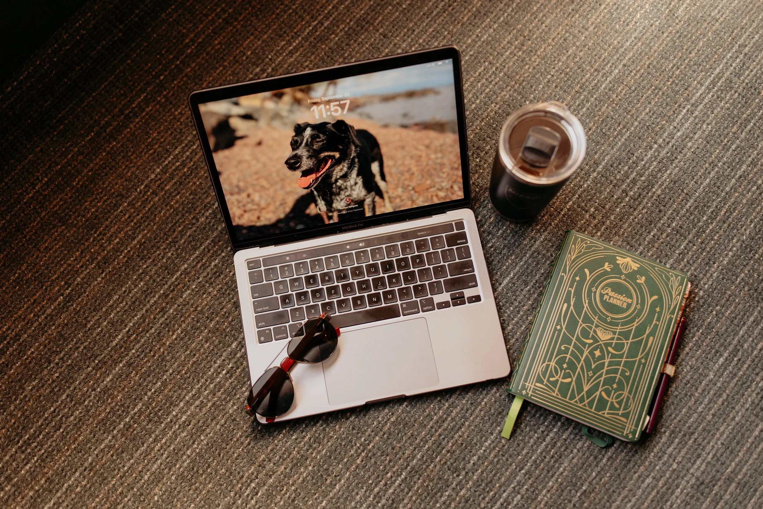 Laptop with a dog wallpaper, sunglasses, green planner, and tumbler on a carpeted floor.