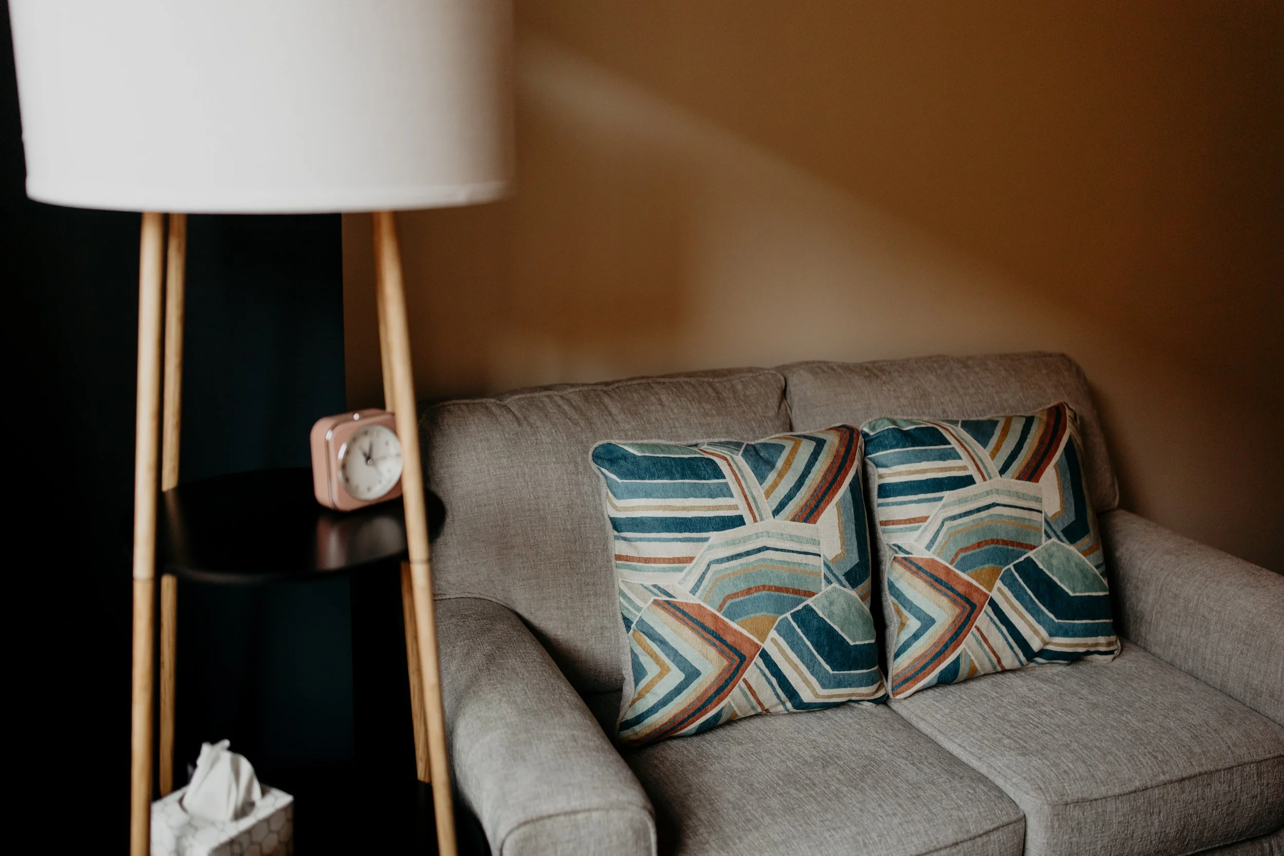 Modern living room with a grey sofa, colorful geometric pillows, and a wooden floor lamp with a white shade. A small clock and tissue box are on the lamp's shelf.