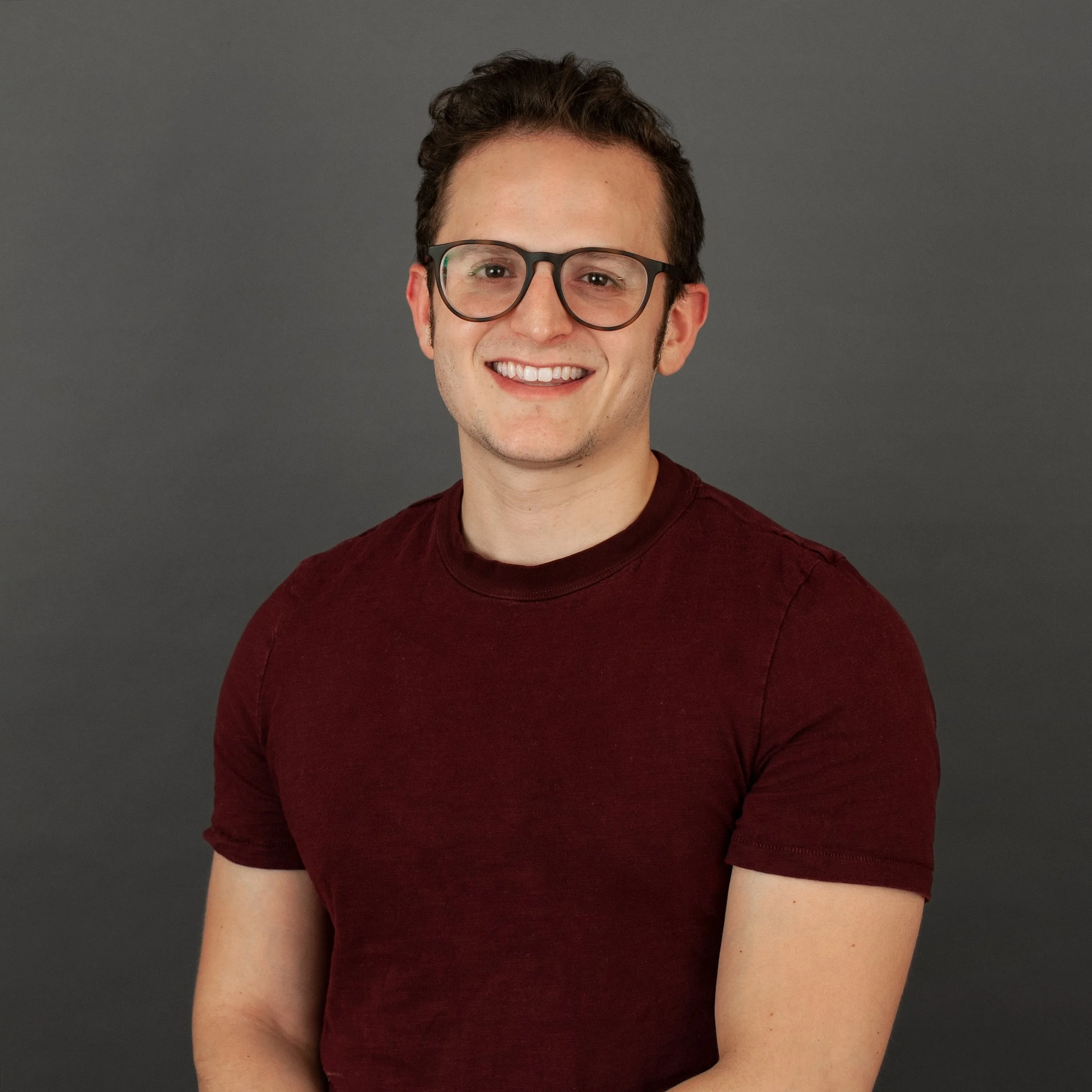 A young man with curly hair, wearing glasses and a dark red T-shirt, smiling in front of a gray background.