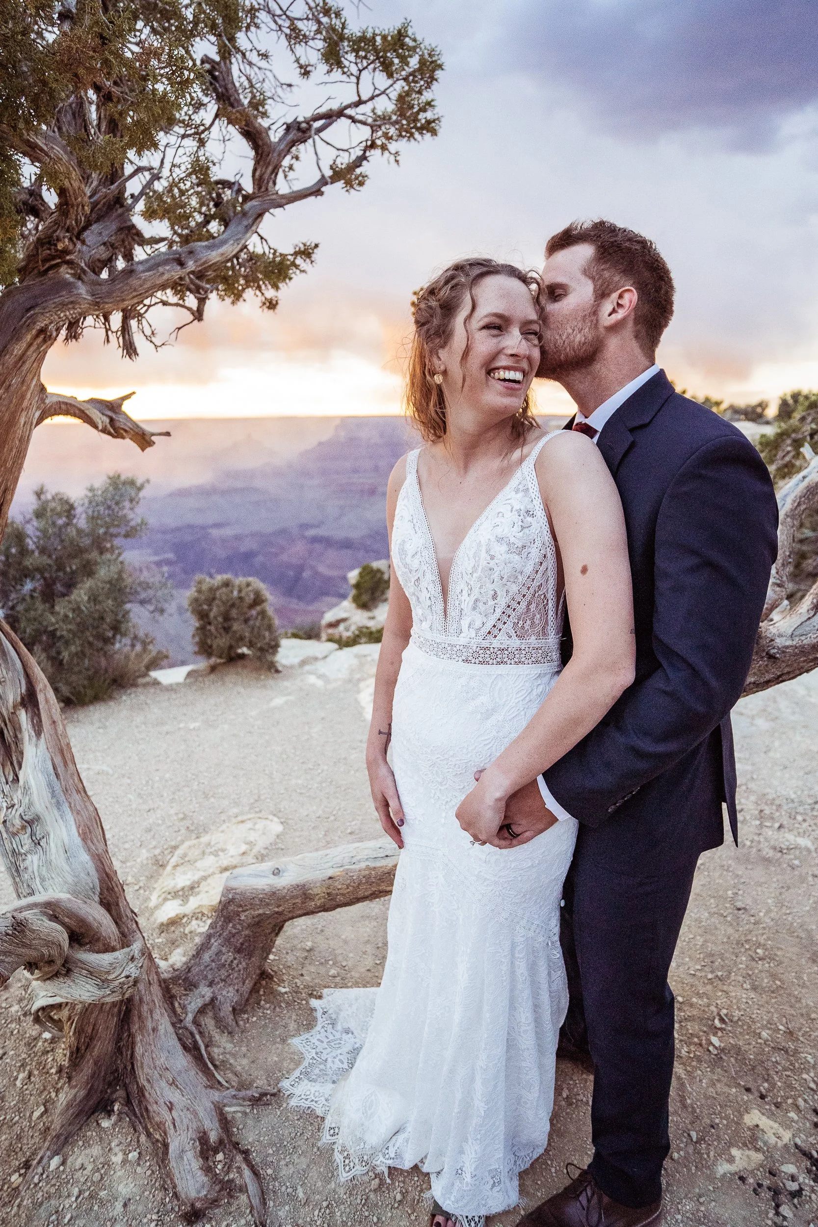 A newlywed couple in wedding attire sharing a joyful moment outdoors near the Grand Canyon at sunset. The woman wears a white lace dress, and the man wears a black suit, with the woman smiling and the man kissing her cheek.