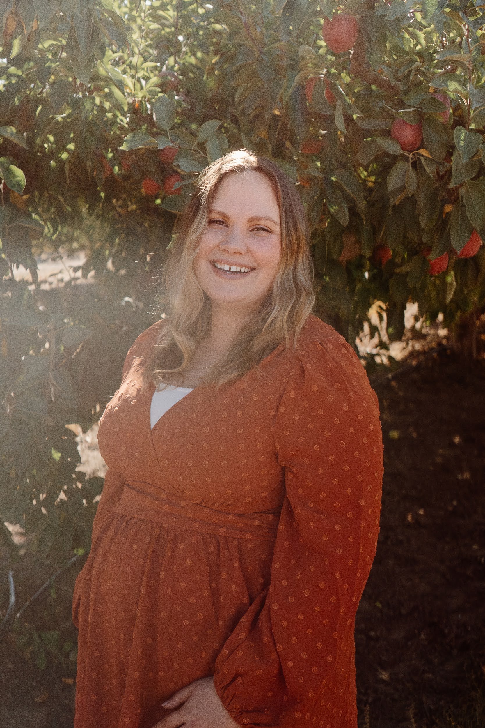 A young woman with wavy blonde hair smiling outdoors, standing in front of a tree with red apples, wearing a rust-colored dress with small patterns.