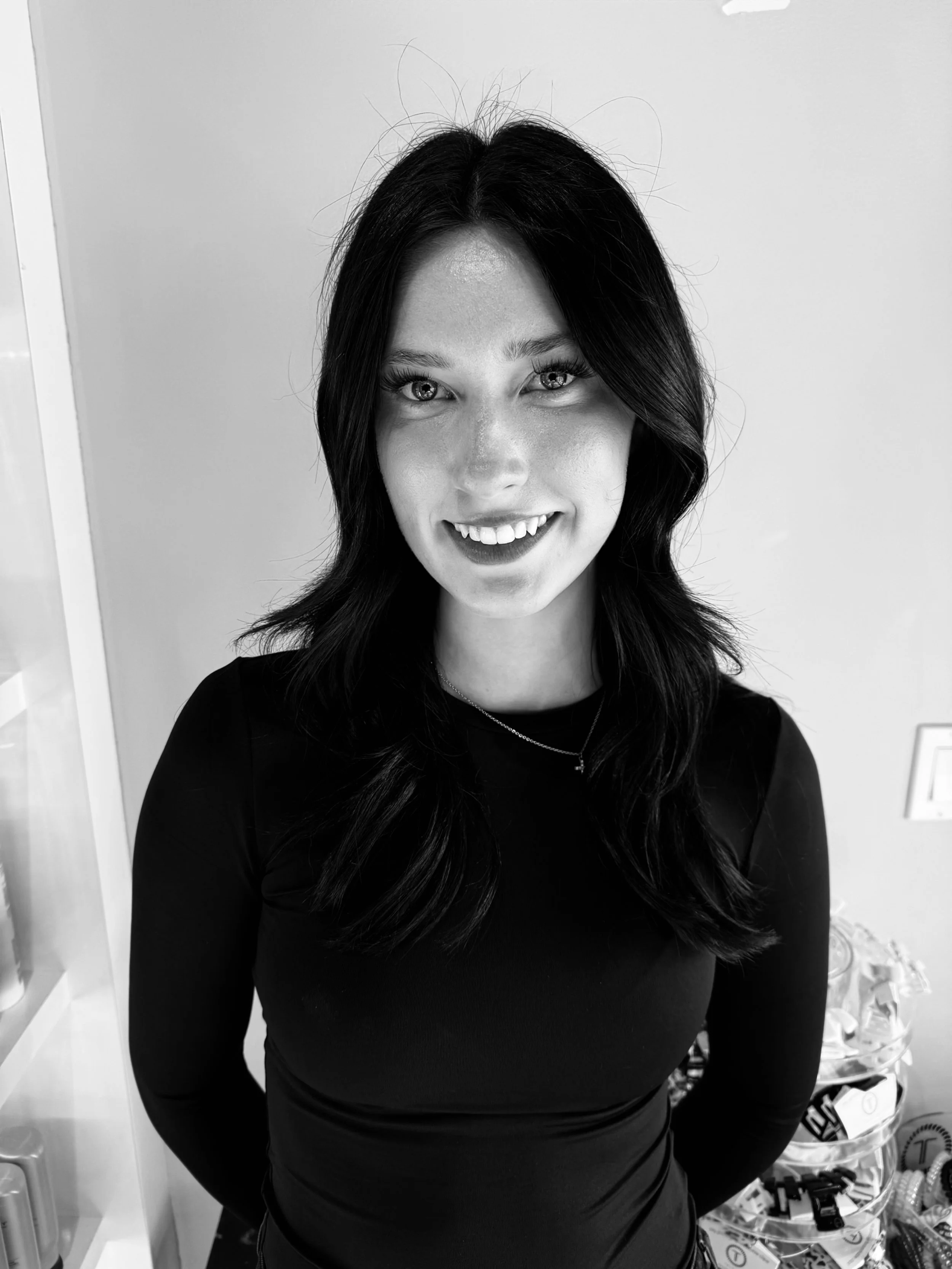 A woman with wavy dark hair smiling at the camera, wearing a long-sleeve black top and a necklace, standing in a room with a neutral wall background.