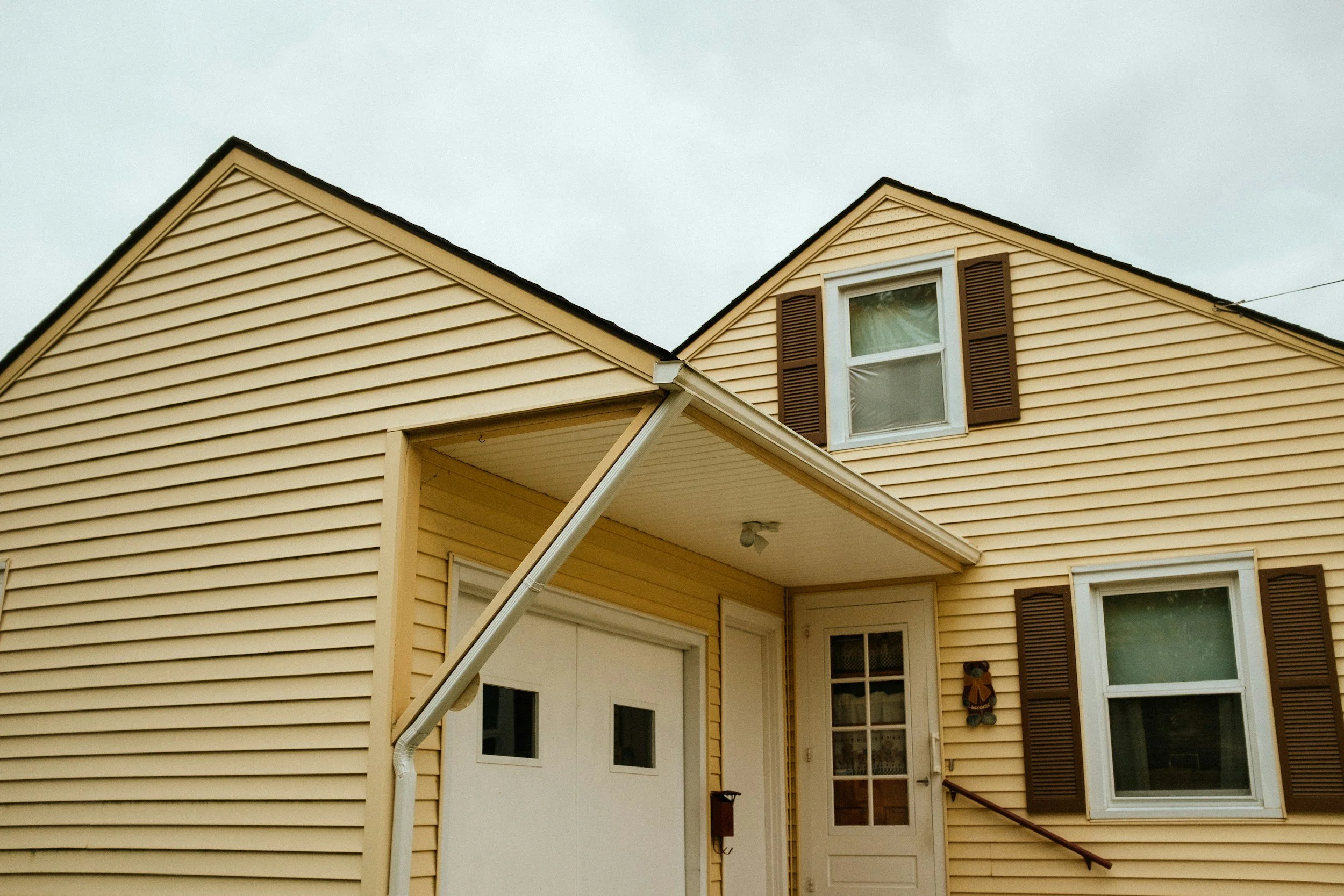 A yellow two-story house with brown shutters, is showing the front entrance and windows, an overcast sky.