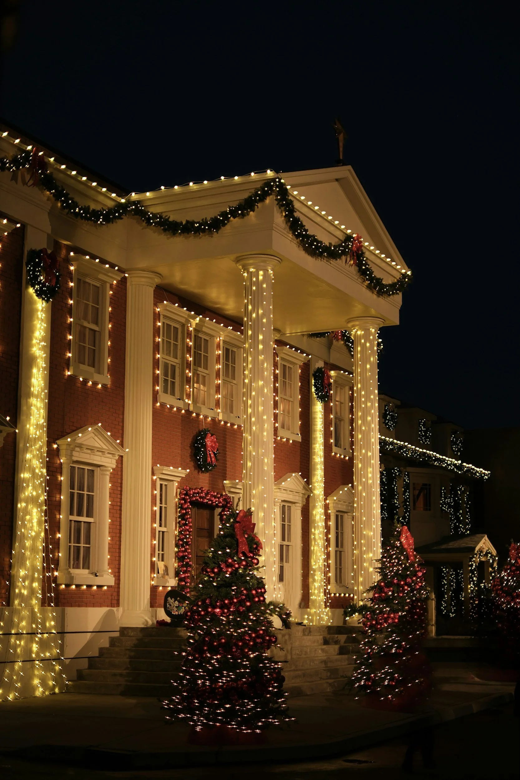 A large house decorated for Christmas with string lights, wreaths, and Christmas trees, illuminated at night.